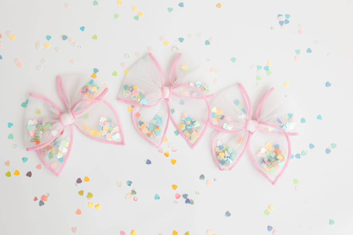 A small, pastel pink mesh shaker bow filled with colorful confetti hearts.