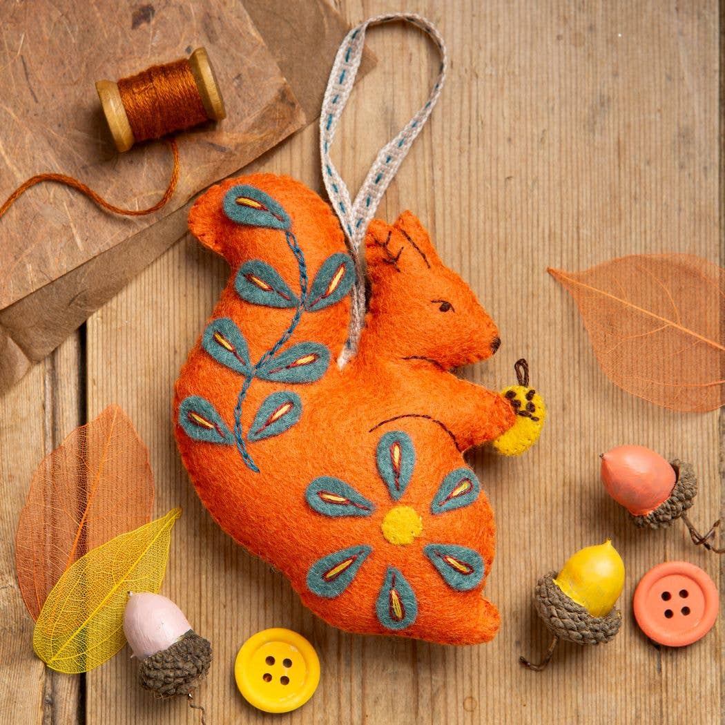 Orange felt squirrel craft with embroidered flowers and holding a yellow acorn.