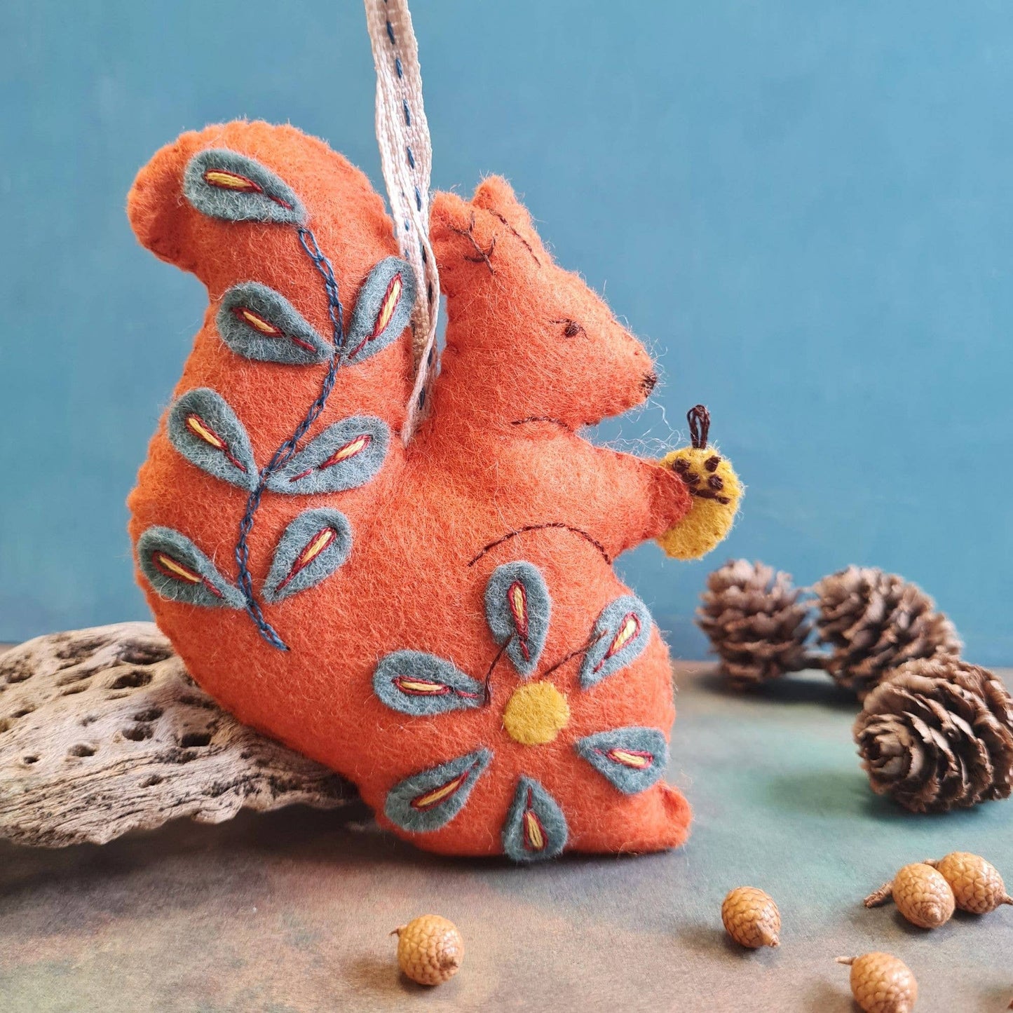 Handmade felt squirrel ornament in orange with embroidered gray leaves and yellow accents, holding a small acorn.