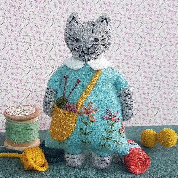 Handmade felt cat in a blue dress with floral embroidery, holding knitting needles and yarn.
