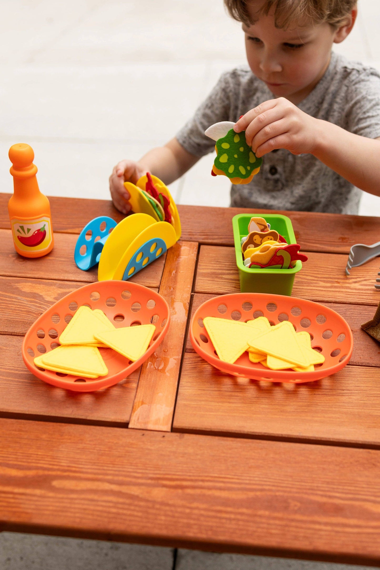 Child assembling tacos with felt ingredients, sauce bottle, nacho baskets, and taco holders on a wooden table.