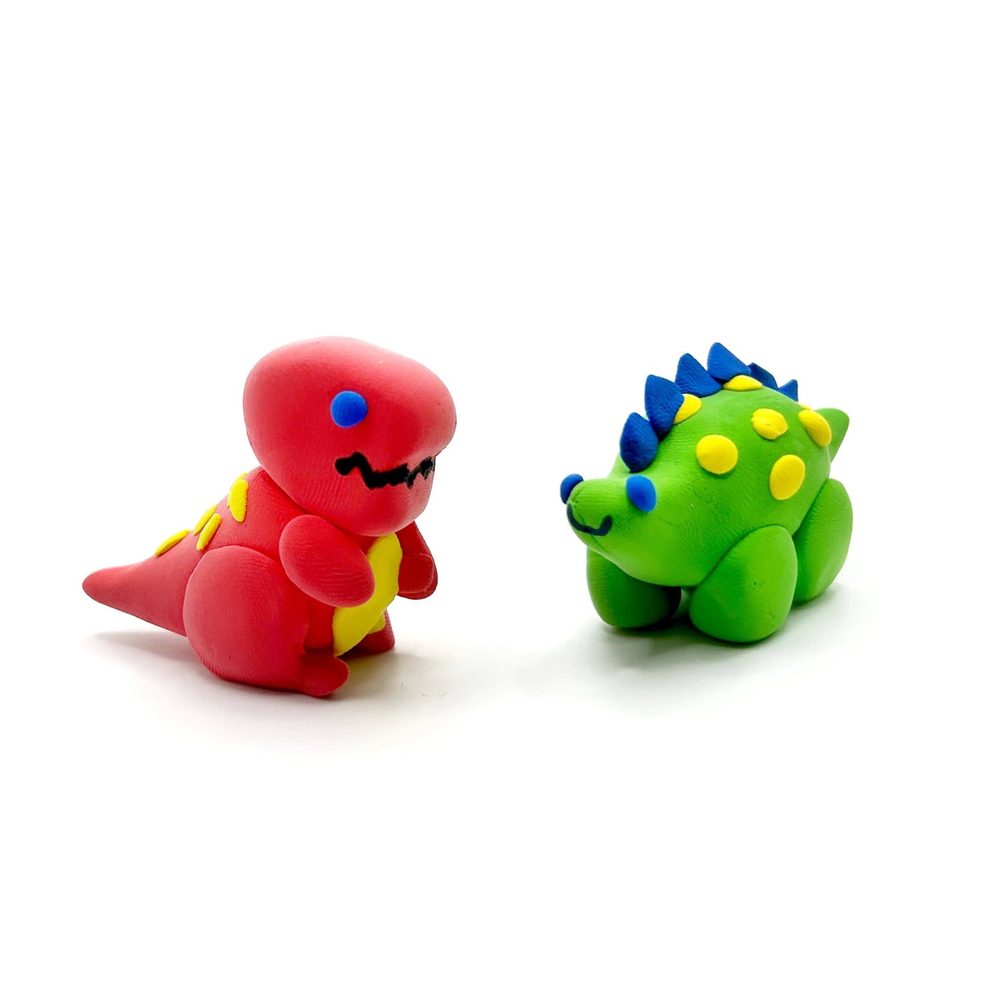 Two colorful air-dry clay dinosaur figurines, one red with yellow spots and one green with blue spikes.