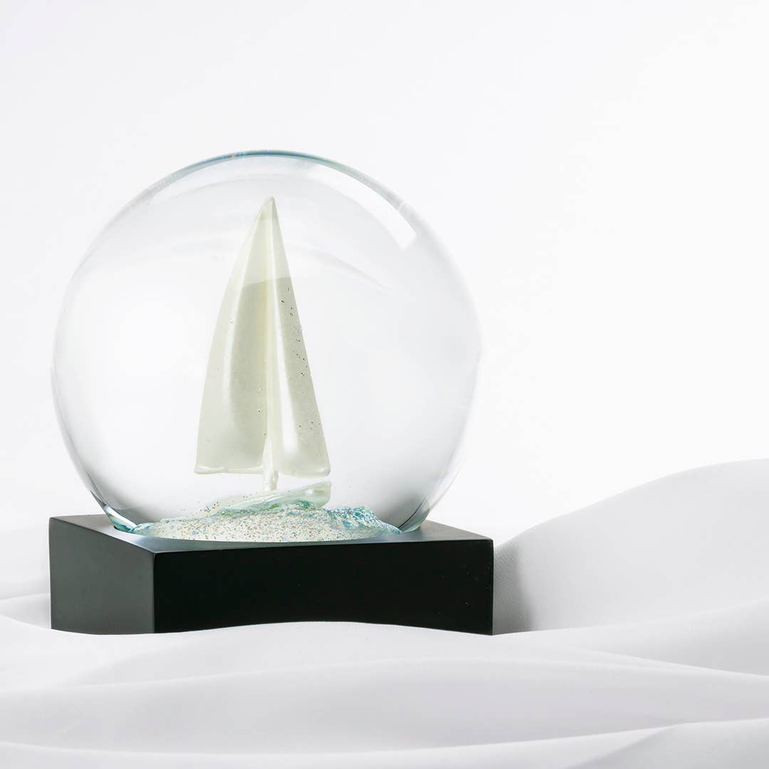 Glass snow globe with a sailboat inside, set on a sleek black base.