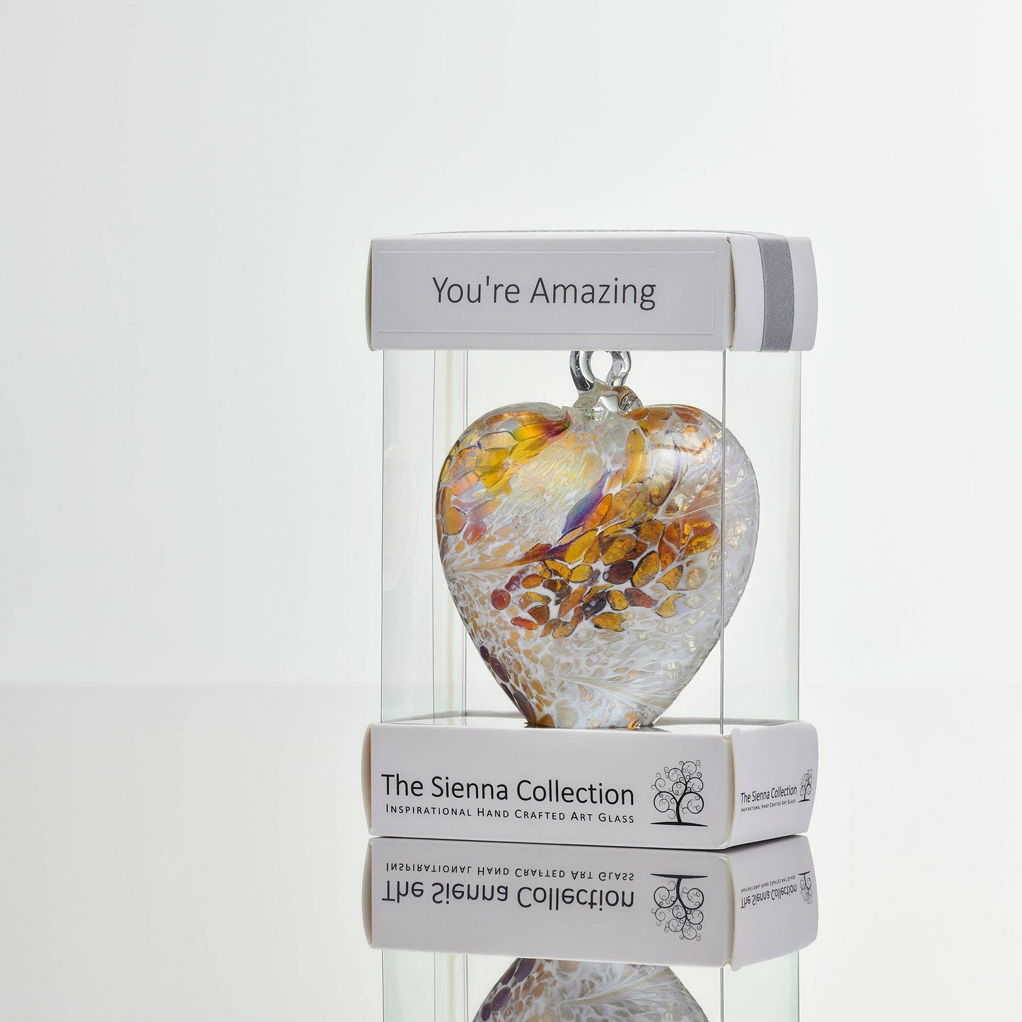 Heart-shaped glass ornament in soft swirls of pastel gold, white, and warm amber tones, displayed in a clear box with the label “You're Amazing.”
