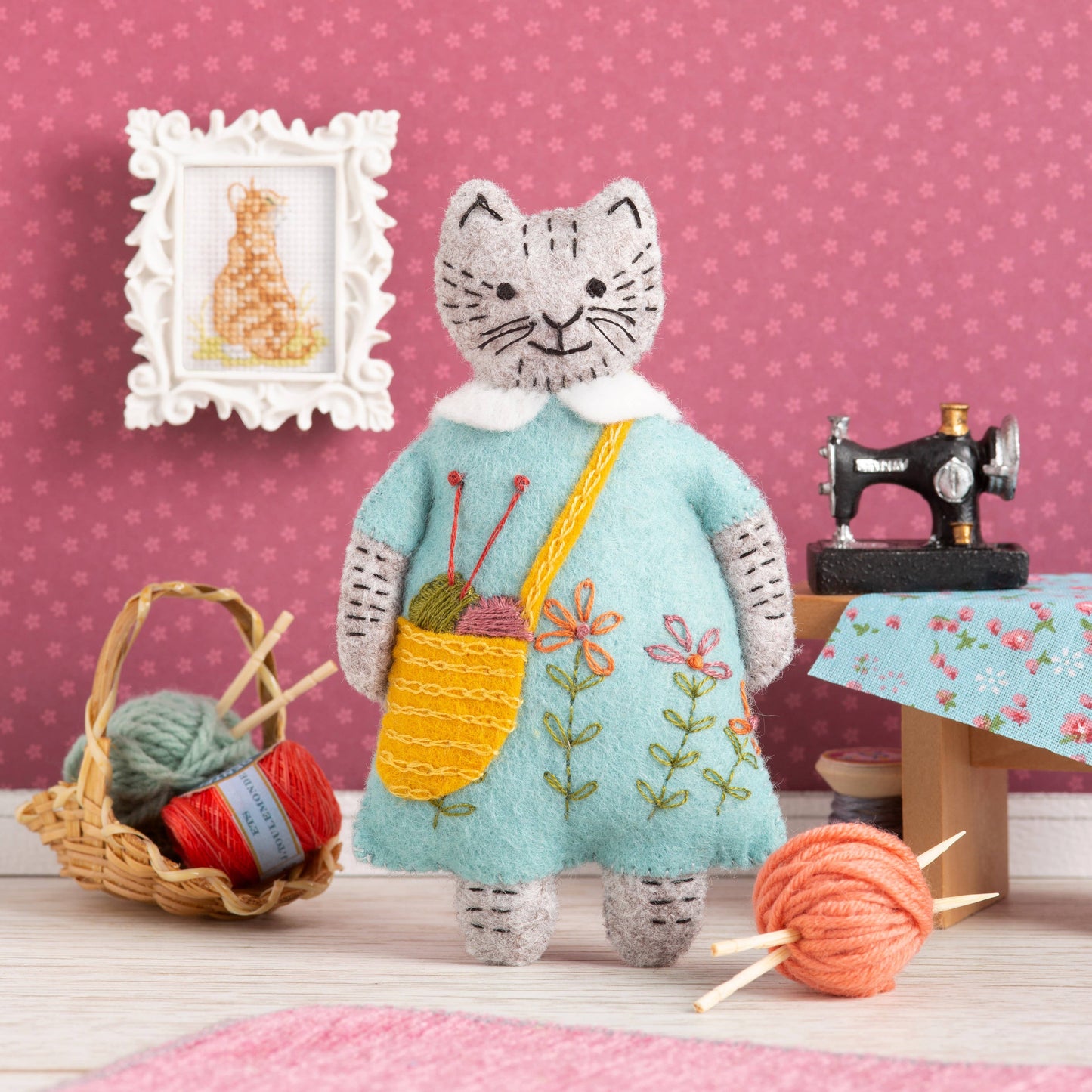 Handmade felt cat in a blue dress with floral embroidery, carrying a yellow bag with yarn and knitting needles.