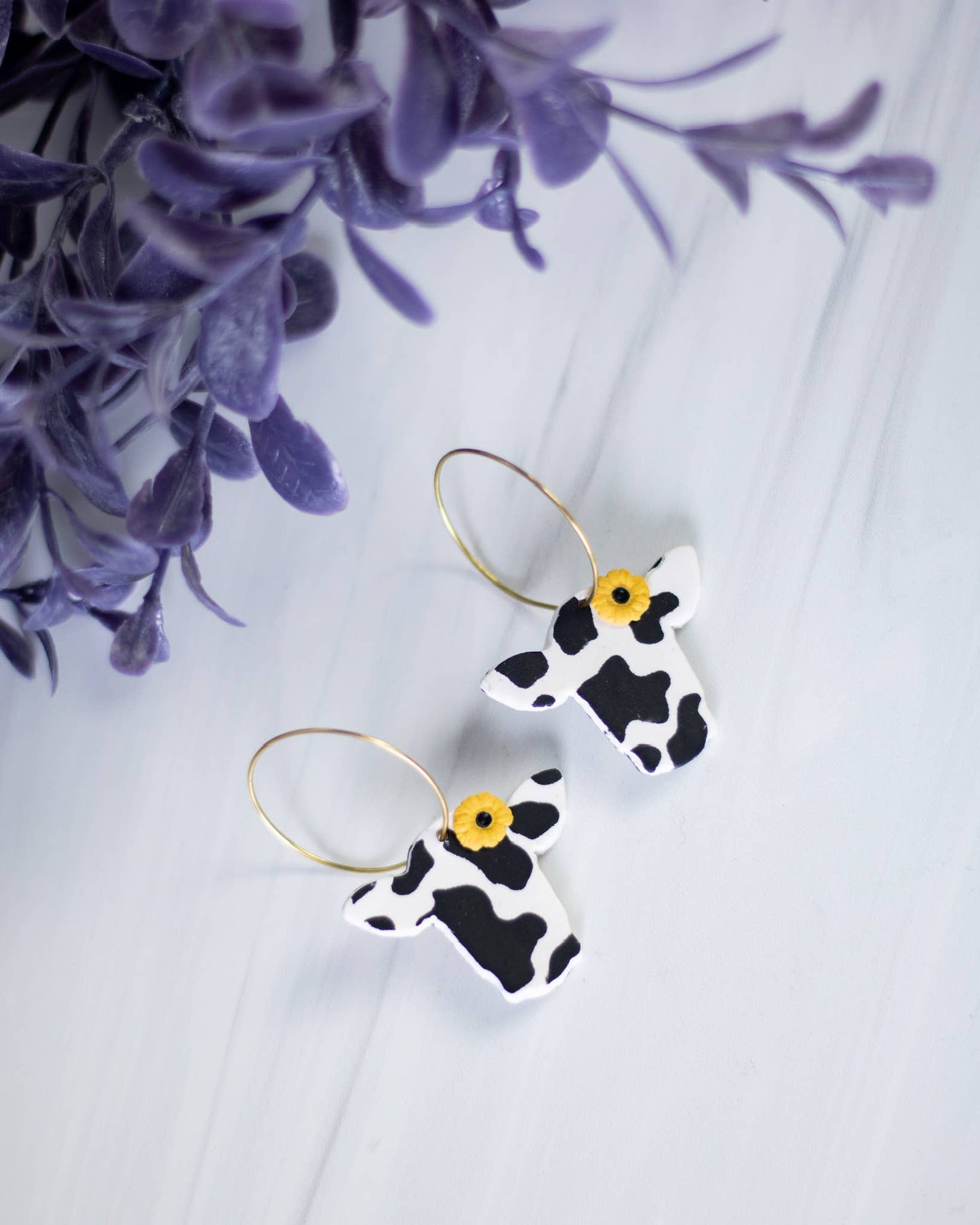 Cow print clay dangle earrings with gold hoops and sunflower studs, perfect for a unique gift.