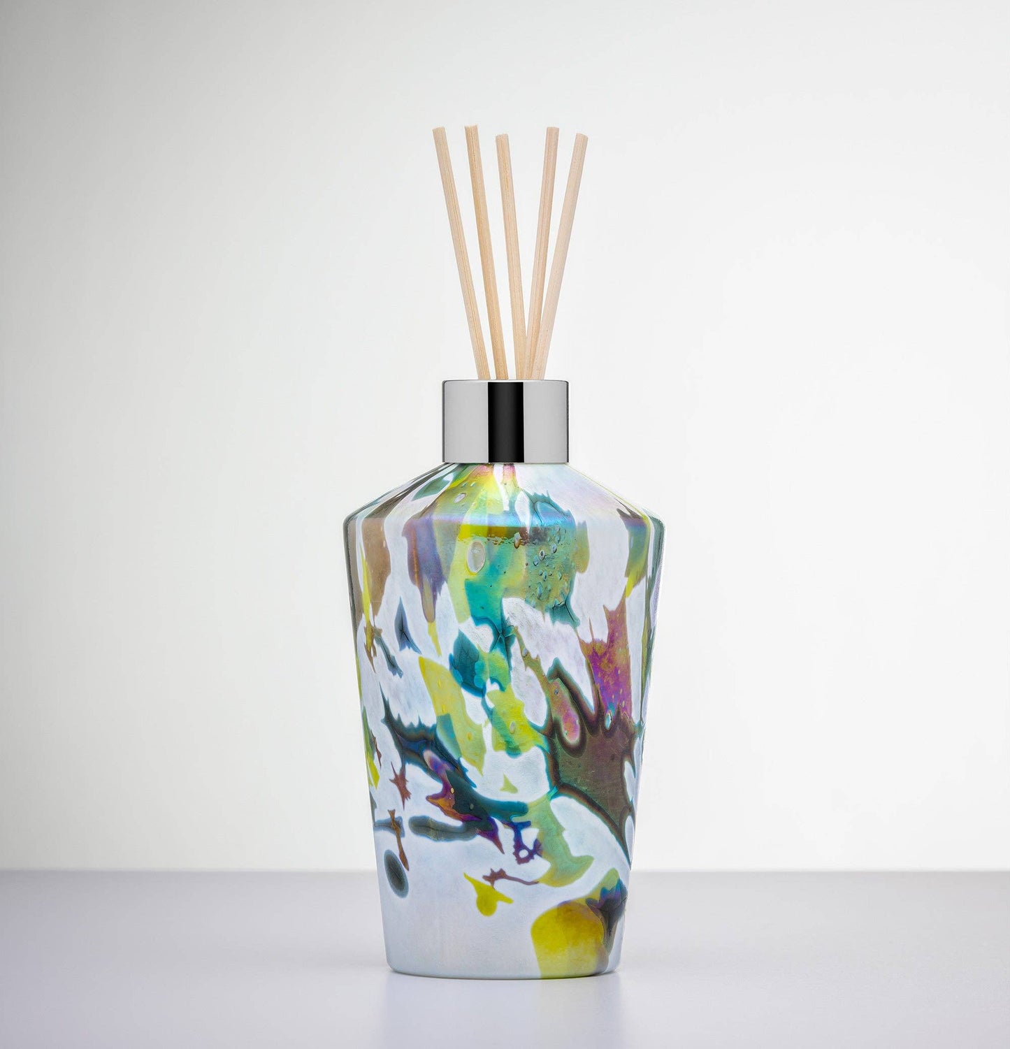 Elegant glass reed diffuser bottle with an abstract swirl of green, yellow, white, and earthy tones; topped with a silver collar and five natural reeds.