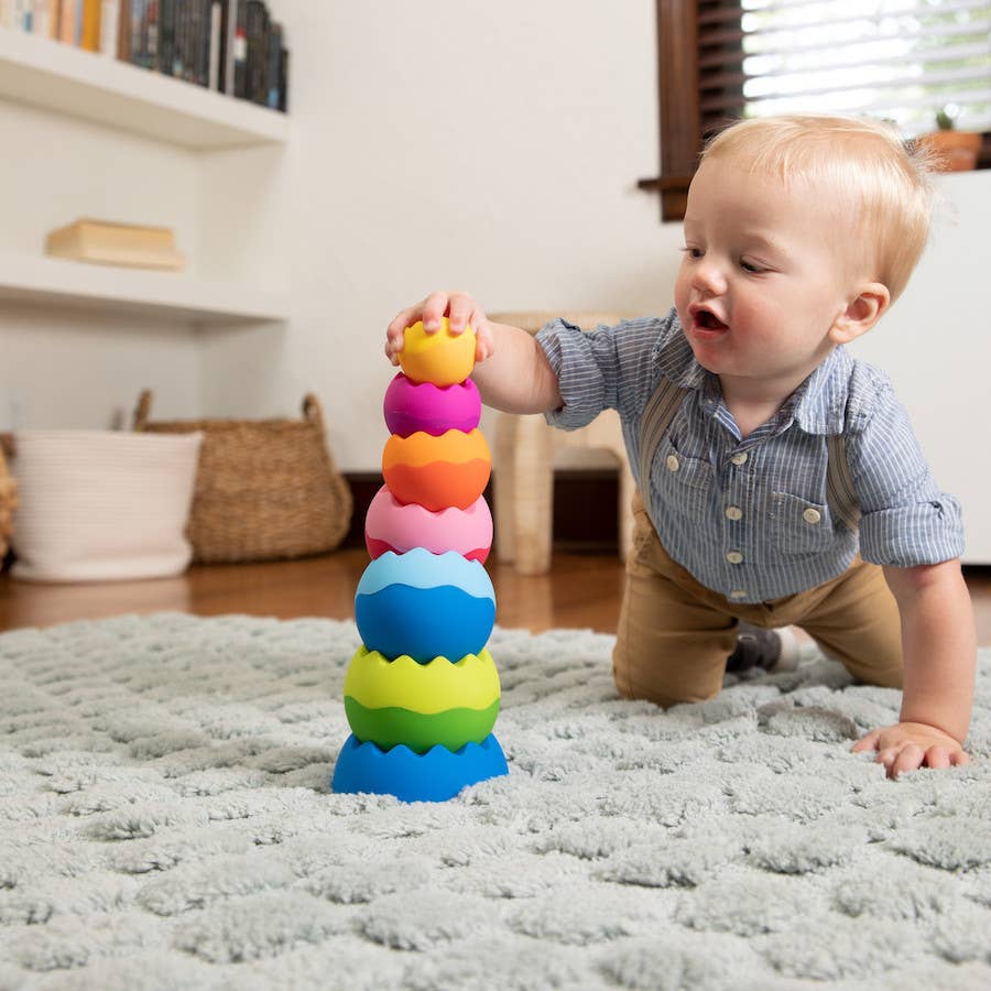 Baby stacking colorful, textured wobble pieces into a tall tower on a soft rug.