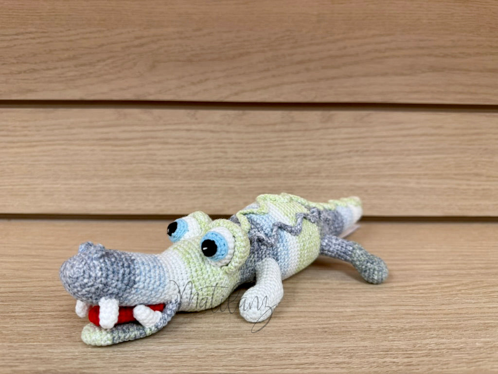 A handmade crochet crocodile toy with blue eyes, striped green and gray body, and red tongue.