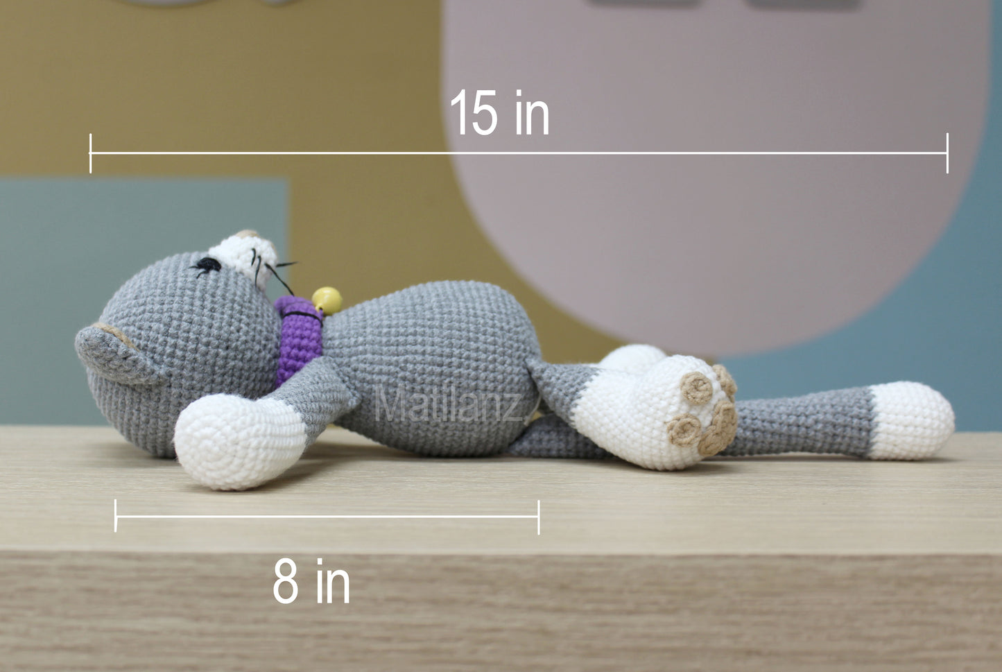 A handmade amigurumi gray cat with white paws and a purple collar, lying on its side.