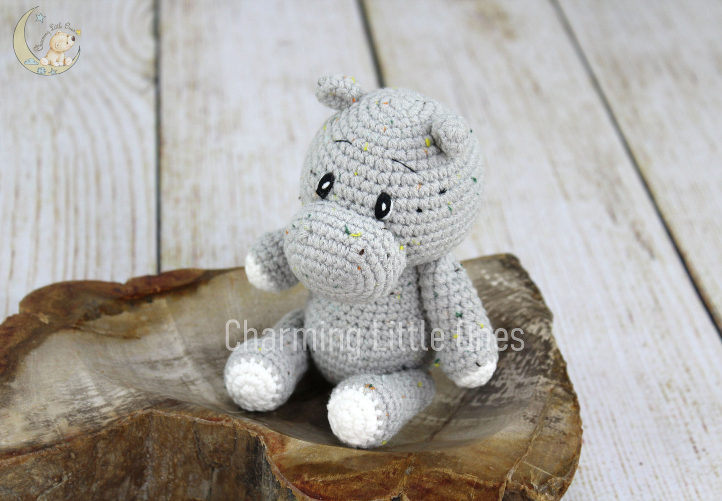A handmade crochet hippo toy with gray and white details, sitting on a wooden slice.