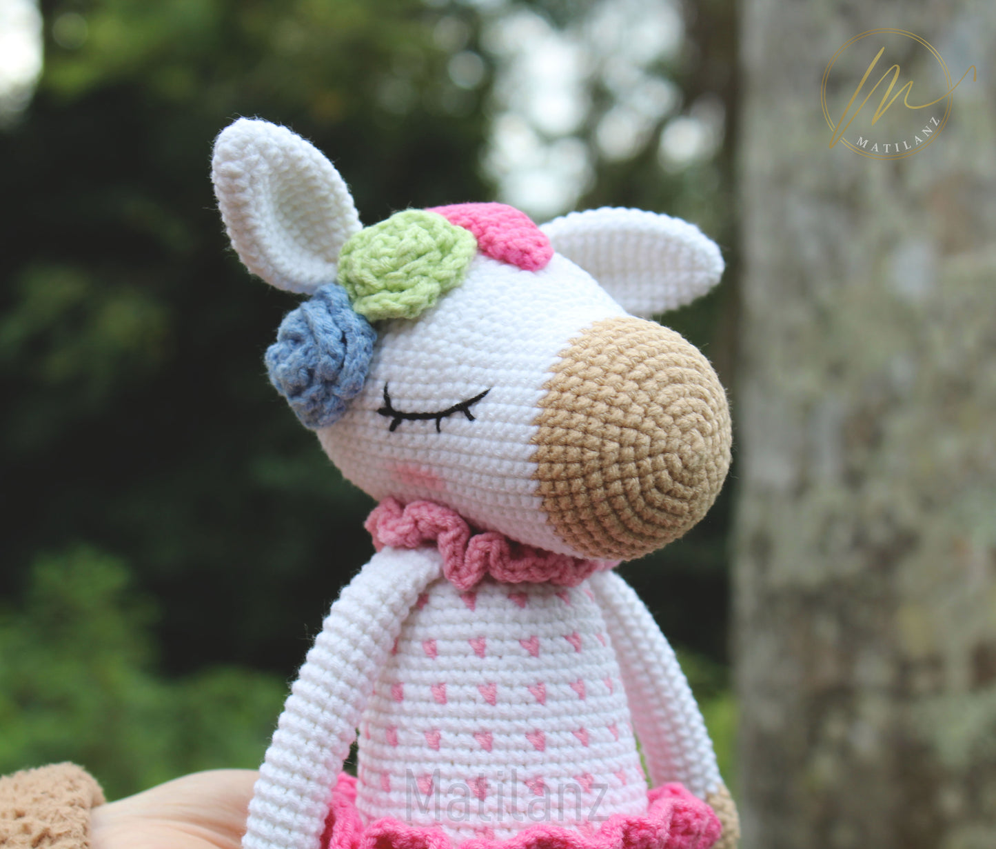 A handmade crochet unicorn with closed eyes, adorned in a pink dress and colorful flower crown.