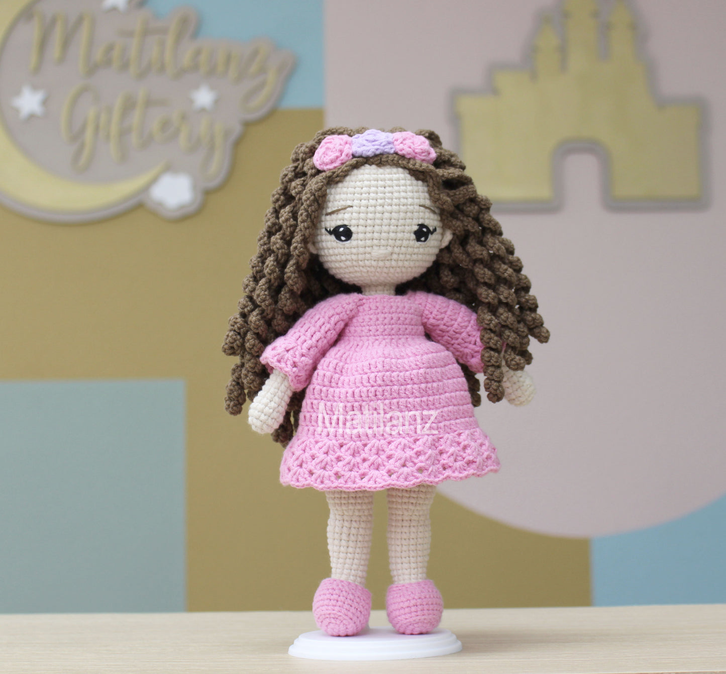 Crochet doll with long brown hair, wearing a pink dress and matching shoes, adorned with a pink and purple headband.