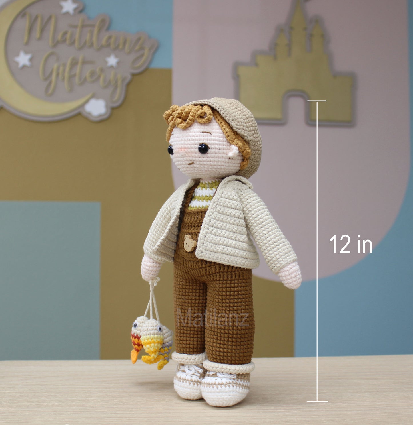 A handmade crochet amigurumi fisherman doll, 12 inches tall, dressed in beige and brown with a hooded jacket and fishing net.