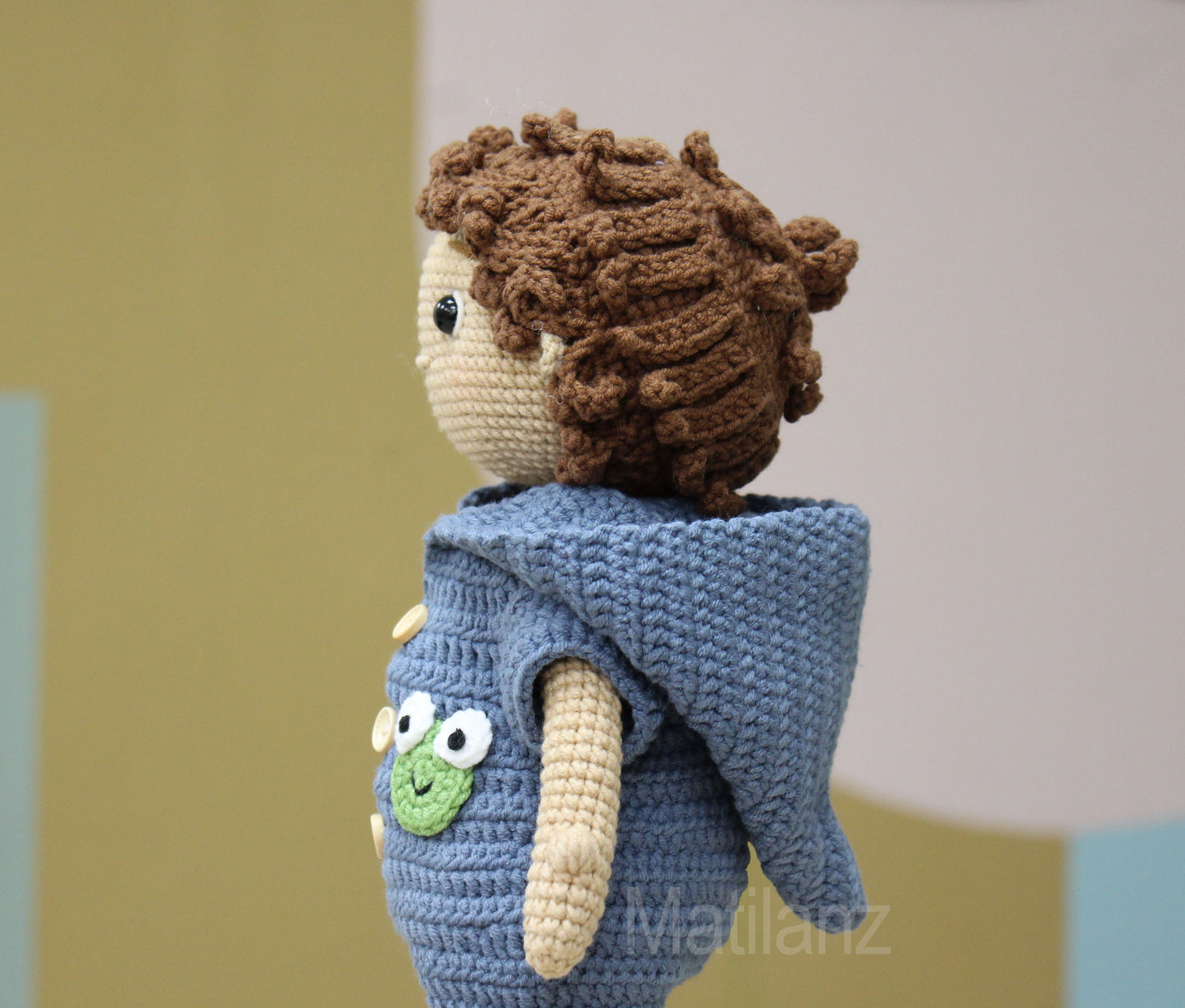 A handmade crochet doll with curly brown hair, wearing a blue outfit featuring a green frog pocket.