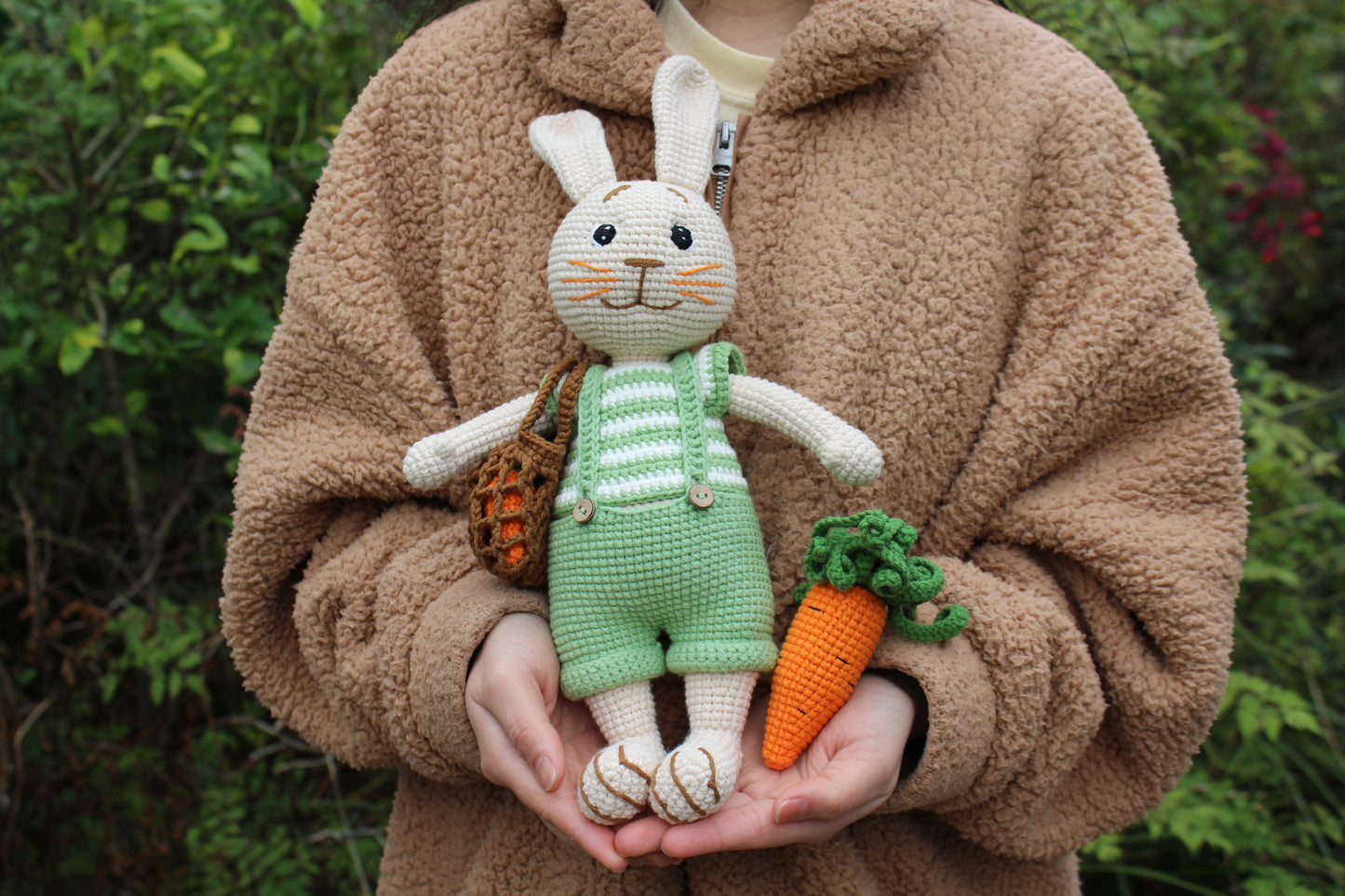 A handmade crochet Easter bunny in green overalls, holding a carrot basket and carrot.