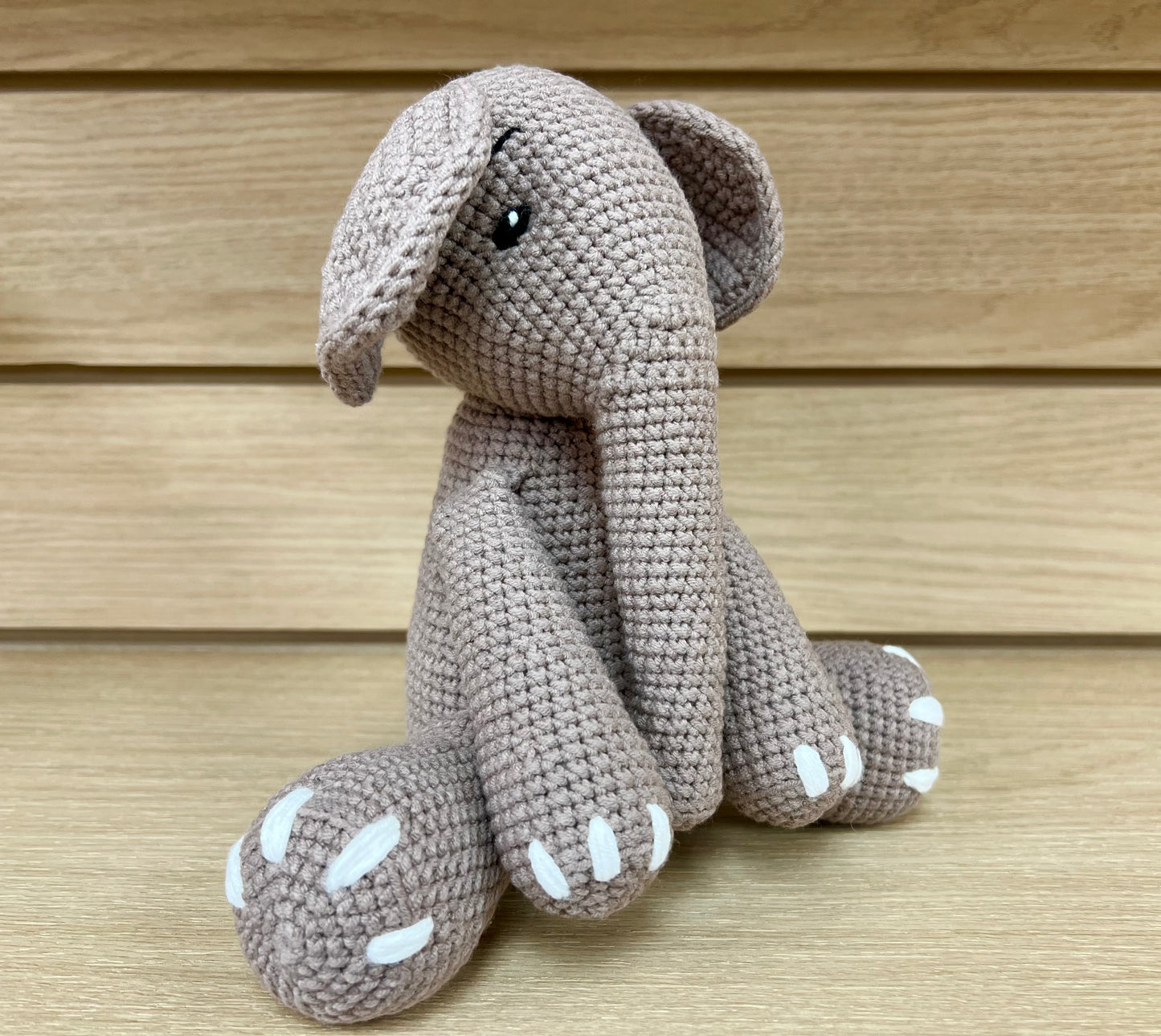 A handmade crochet elephant with large ears and white-tipped paws, sitting on a wooden surface.