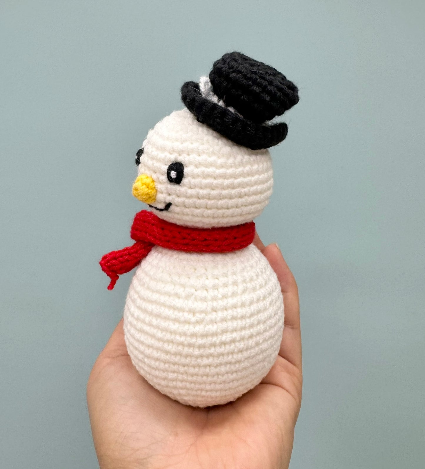 Crochet Christmas snowman with a black top hat, red scarf, and cheerful yellow nose.