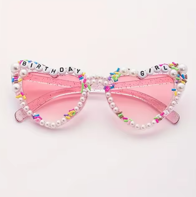 Pink heart-shaped sunglasses decorated with pearl beading, rainbow sprinkles, and the words “Birthday Girl” on the frames.