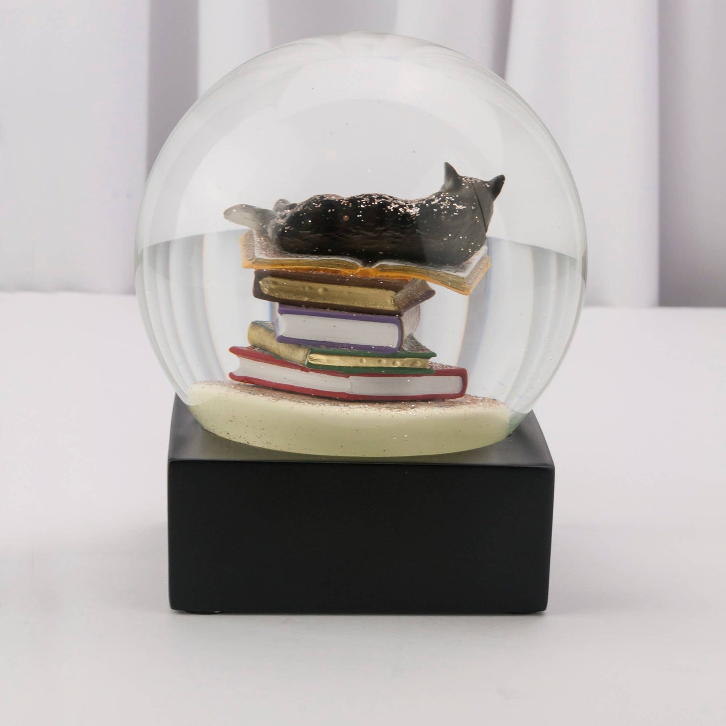 A charming snow globe featuring a black cat resting on colorful books, adorned with a gold butterfly detail.
