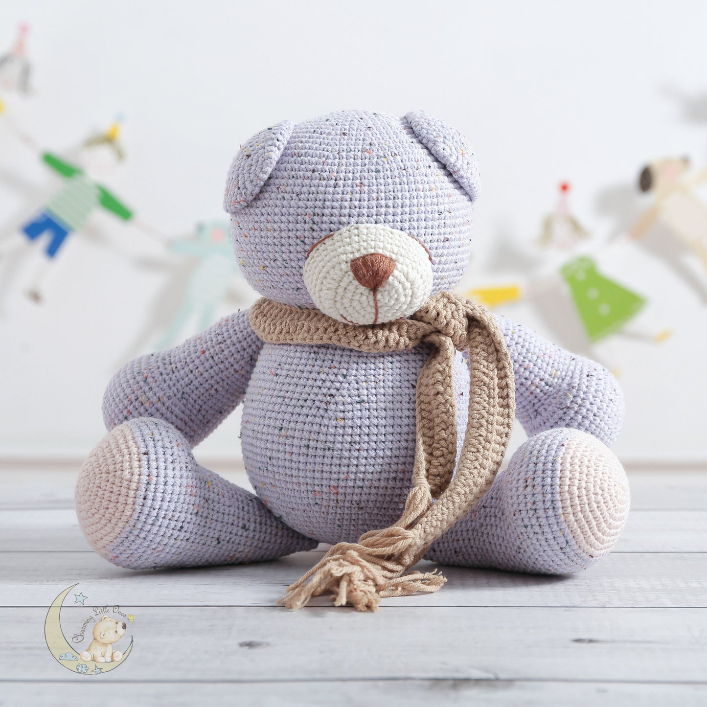 A handmade crocheted teddy bear in light purple with a beige scarf, sitting on a wooden floor.