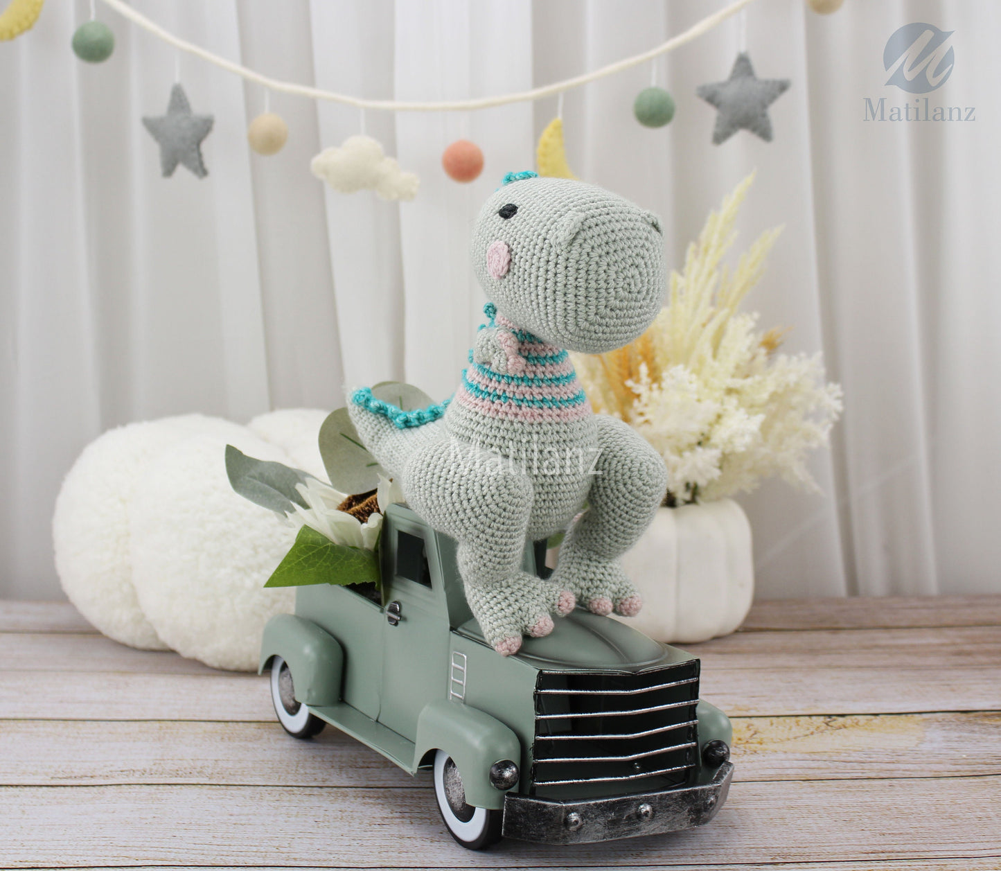 A handmade crochet TRex dinosaur toy with pastel colors and a striped scarf, sitting on a green vintage truck.