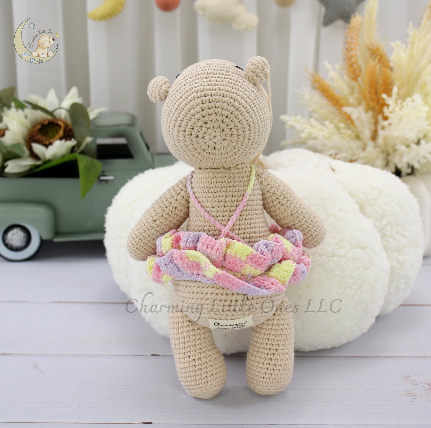 A handmade crochet hippo doll with a pastel checkered skirt, sitting on a white fluffy pillow.