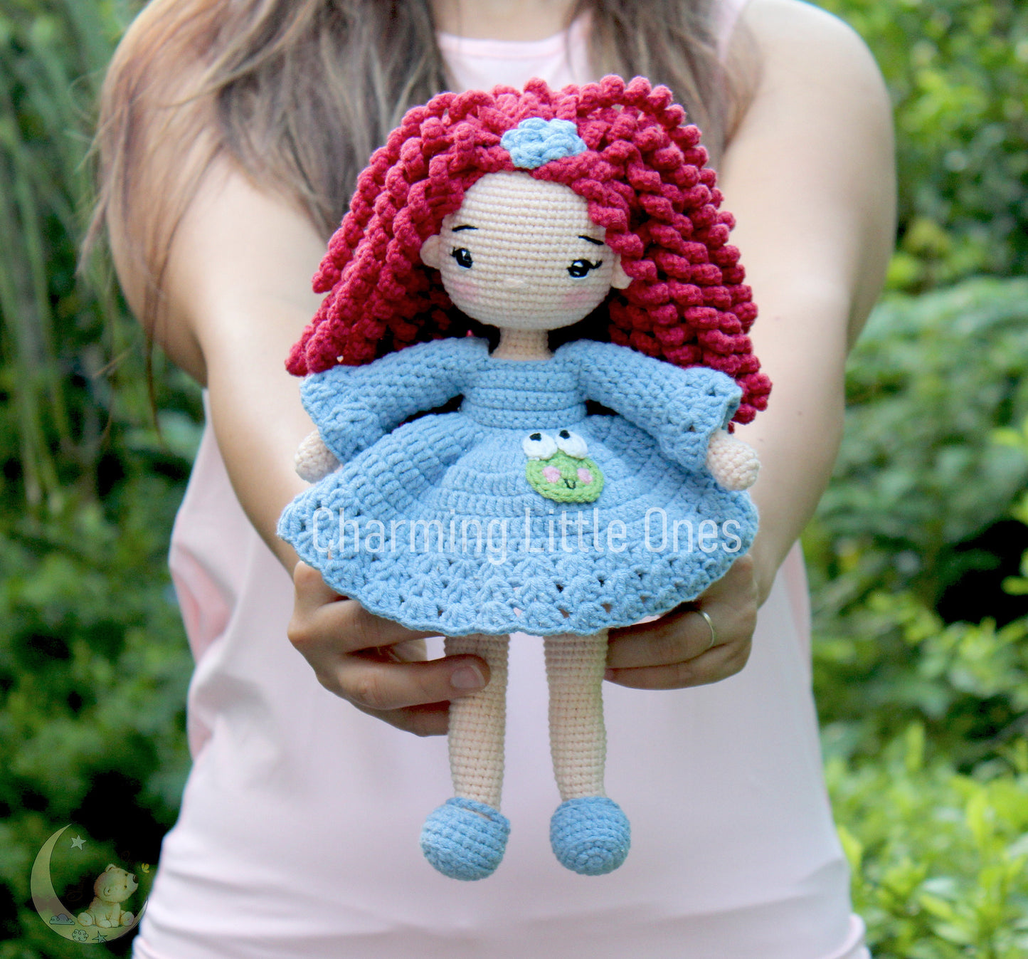 Handmade crochet doll with pink hair, blue dress, and frog appliqué, perfect for cuddling.