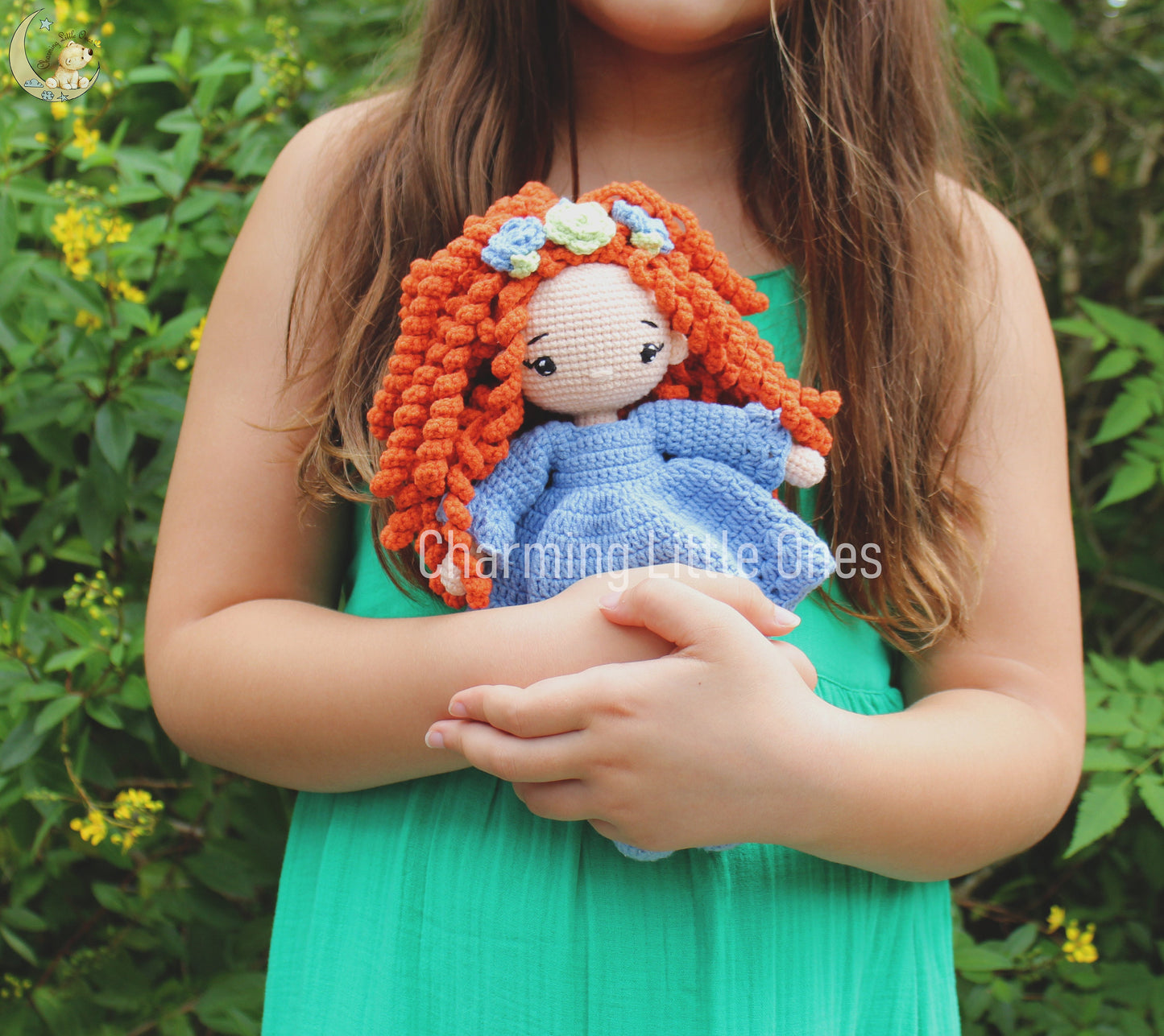 A handmade crochet doll with orange curly hair and a blue dress, adorned with colorful flowers.