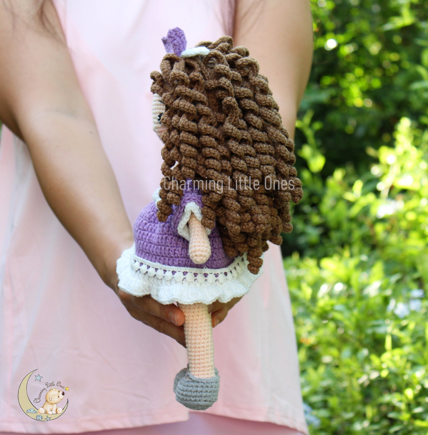 A handmade crochet doll with brown curly hair, purple dress, and white lace trim.