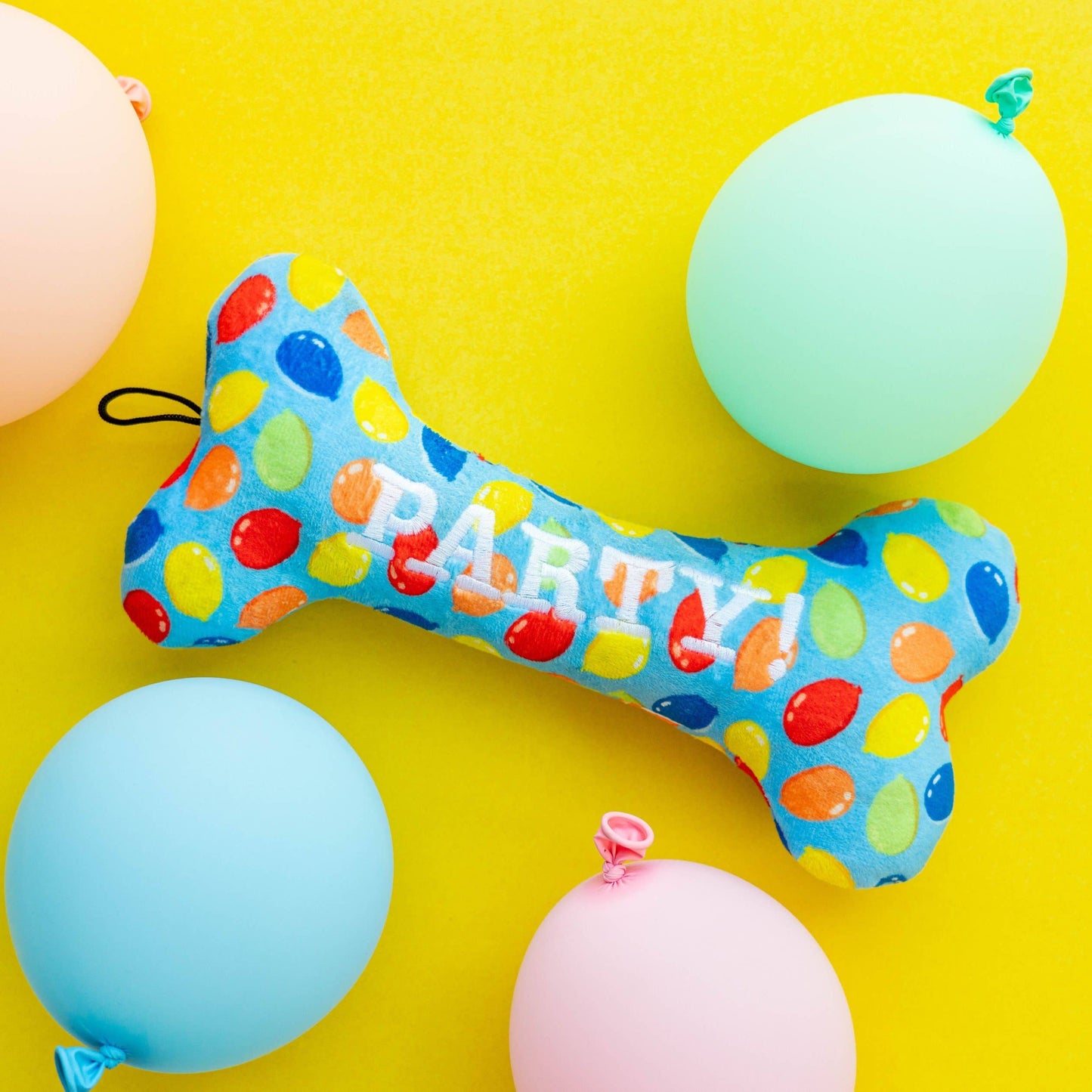 Bone-shaped dog toy in blue with colorful balloon print and "PARTY!" text, surrounded by balloons.