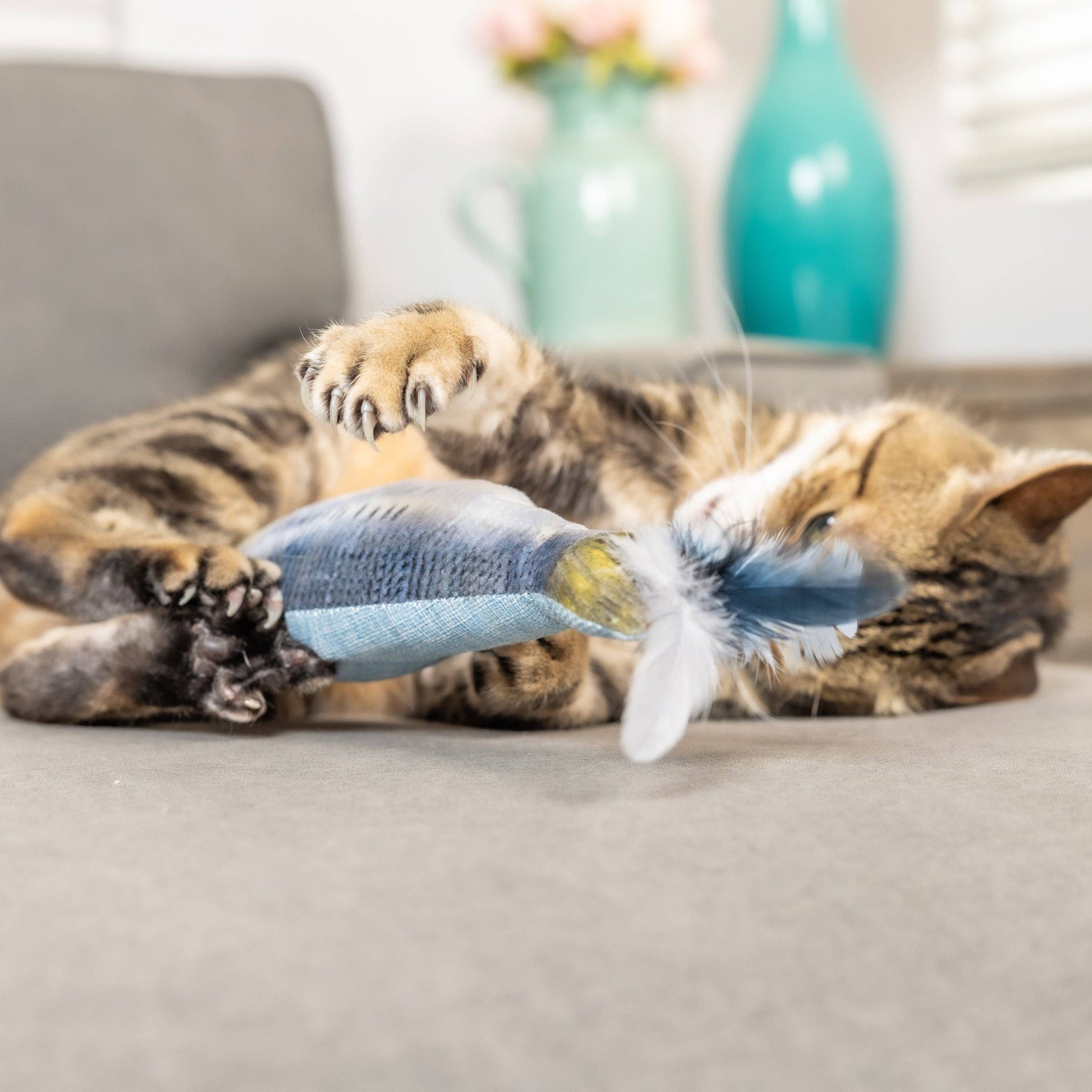 Plush blue fish cat toy with feathers and catnip, being playfully pawed by a cat.