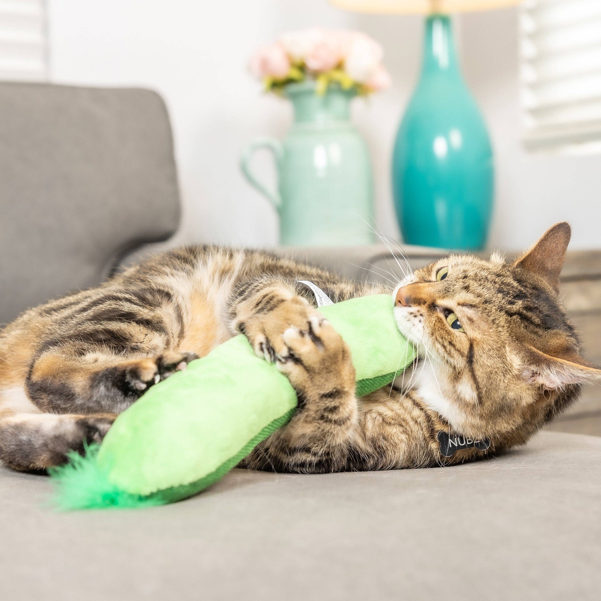 A green crinkle-textured pickle-shaped cat toy with a feathered end, engaging a tabby cat.