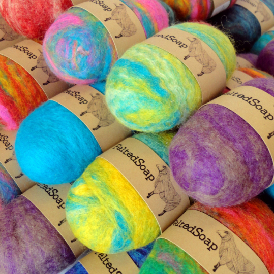A collection of colorful, oval-shaped felted soaps wrapped in kraft paper bands labeled "Felted Soap", featuring vibrant swirls of wool in shades of blue, yellow, purple, red, and pin