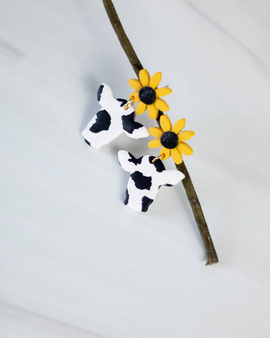 Pair of handcrafted cow-print clay earrings with bright yellow sunflower tops, displayed on a twig for a rustic aesthetic.