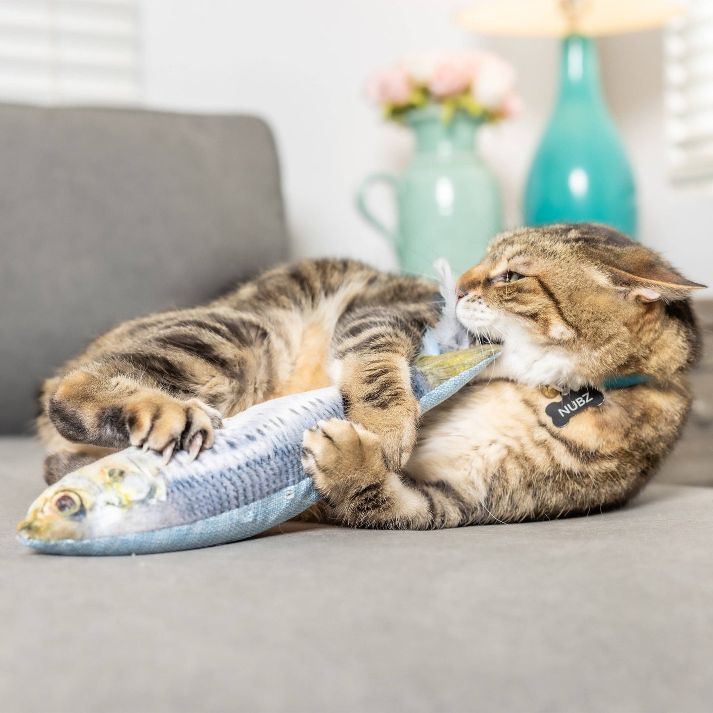 Plush blue fish cat toy with feathers and catnip, being playfully chewed by a tabby cat.