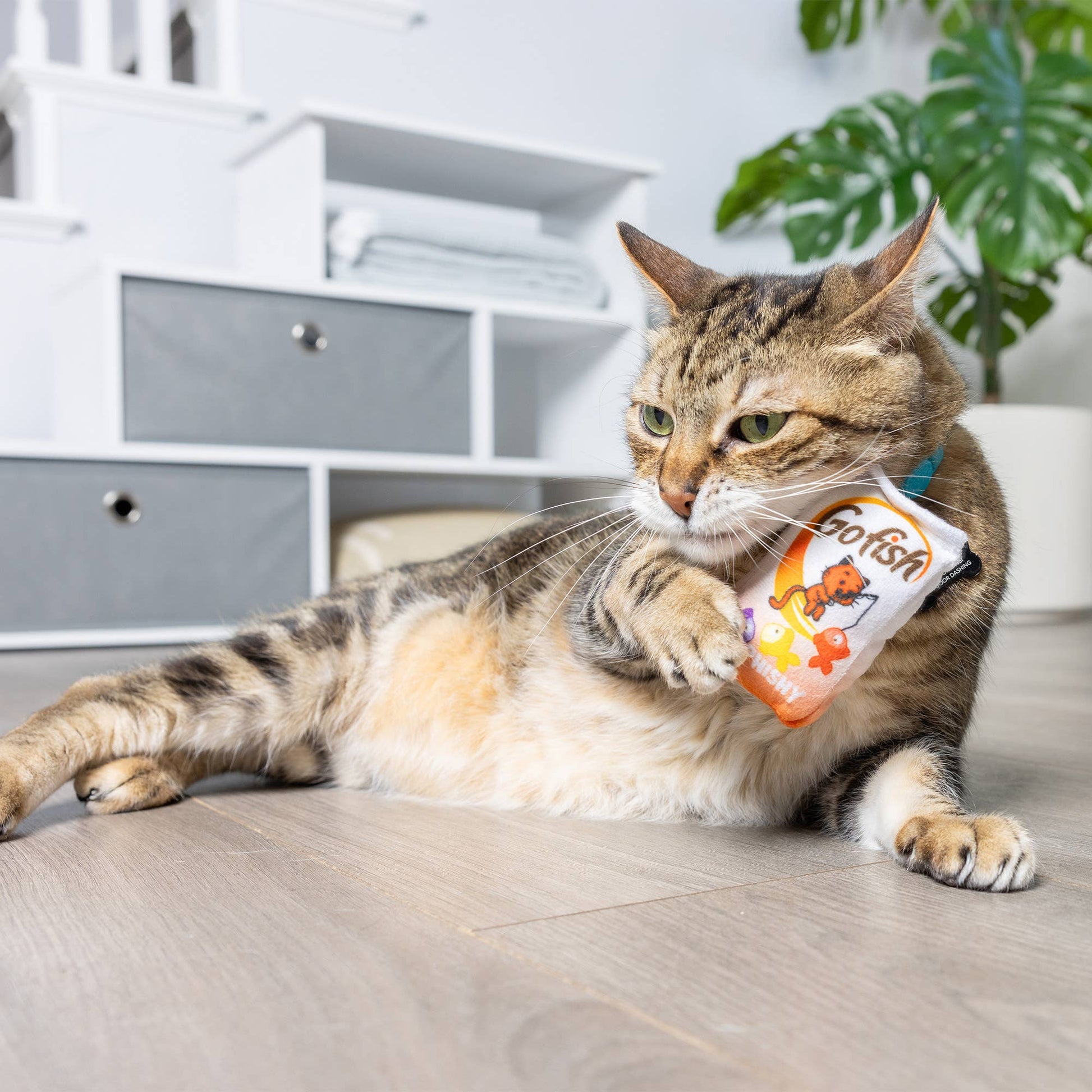 Plush cat toy with "Go Fish" design, featuring catnip and a playful fish图案, held by a relaxed tabby cat.