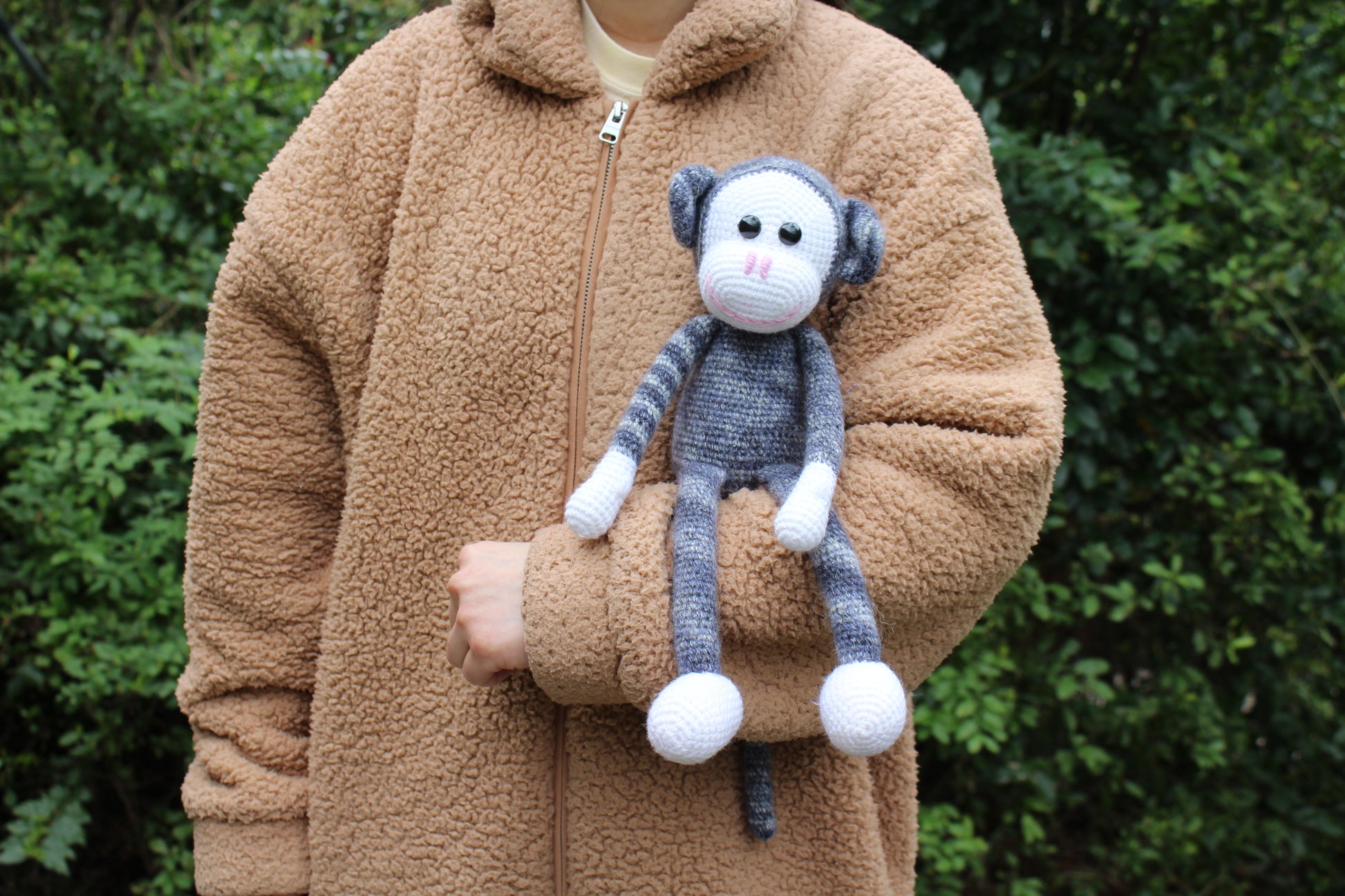 Handmade crochet monkey with gray and white fur, black eyes, and a pink nose, sitting on a sleeve.