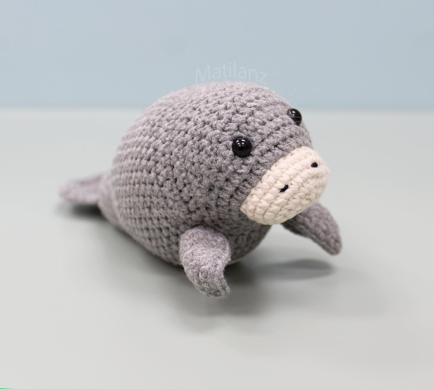 A gray handmade crochet manatee plush with a cream-colored snout and black eyes, posed on a light surface.