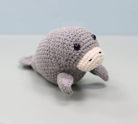 A gray handmade crochet manatee plush with a cream-colored snout and black eyes, posed on a light surface.
