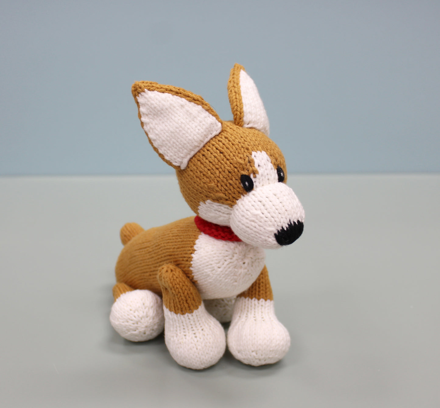 Handmade knitted corgi plush with brown and white fur, a red collar, and perky ears.