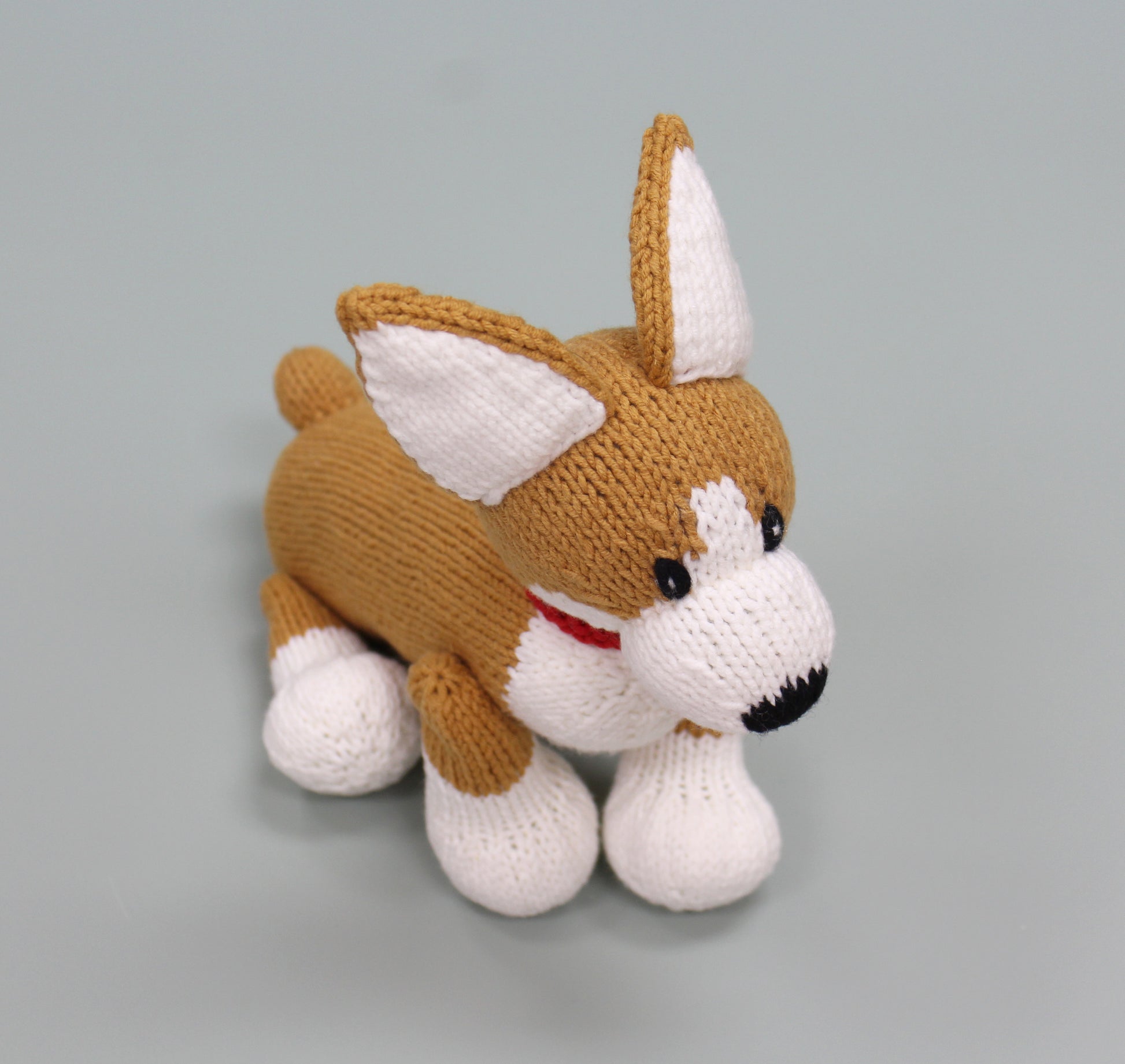 Handmade knitted stuffed animal resembling a corgi with brown and white fur and a red collar.