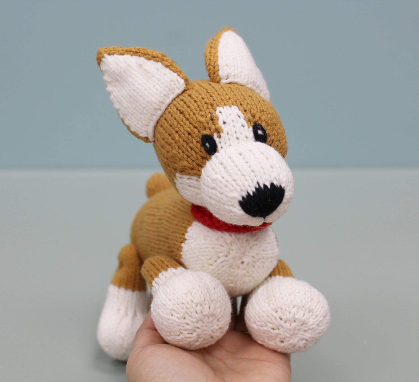 Brown and white handmade knitted corgi plush toy with a red collar, held in hand.