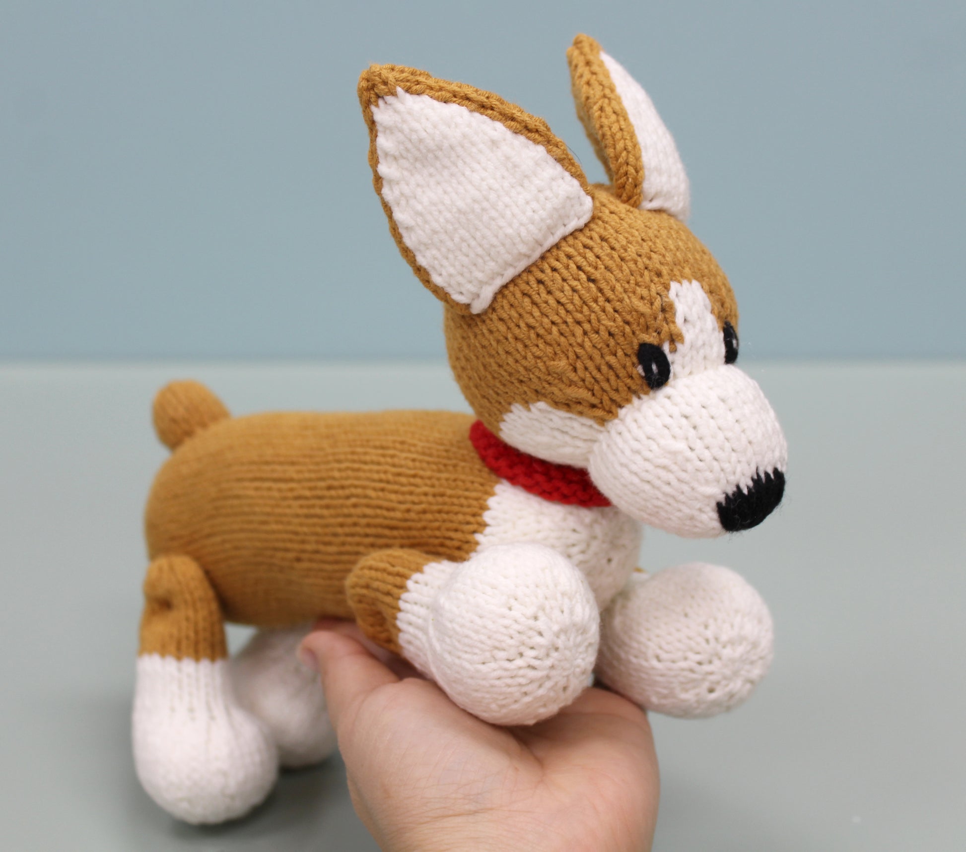 Brown and white handmade knitted corgi plush toy with a red collar, held gently in hand.