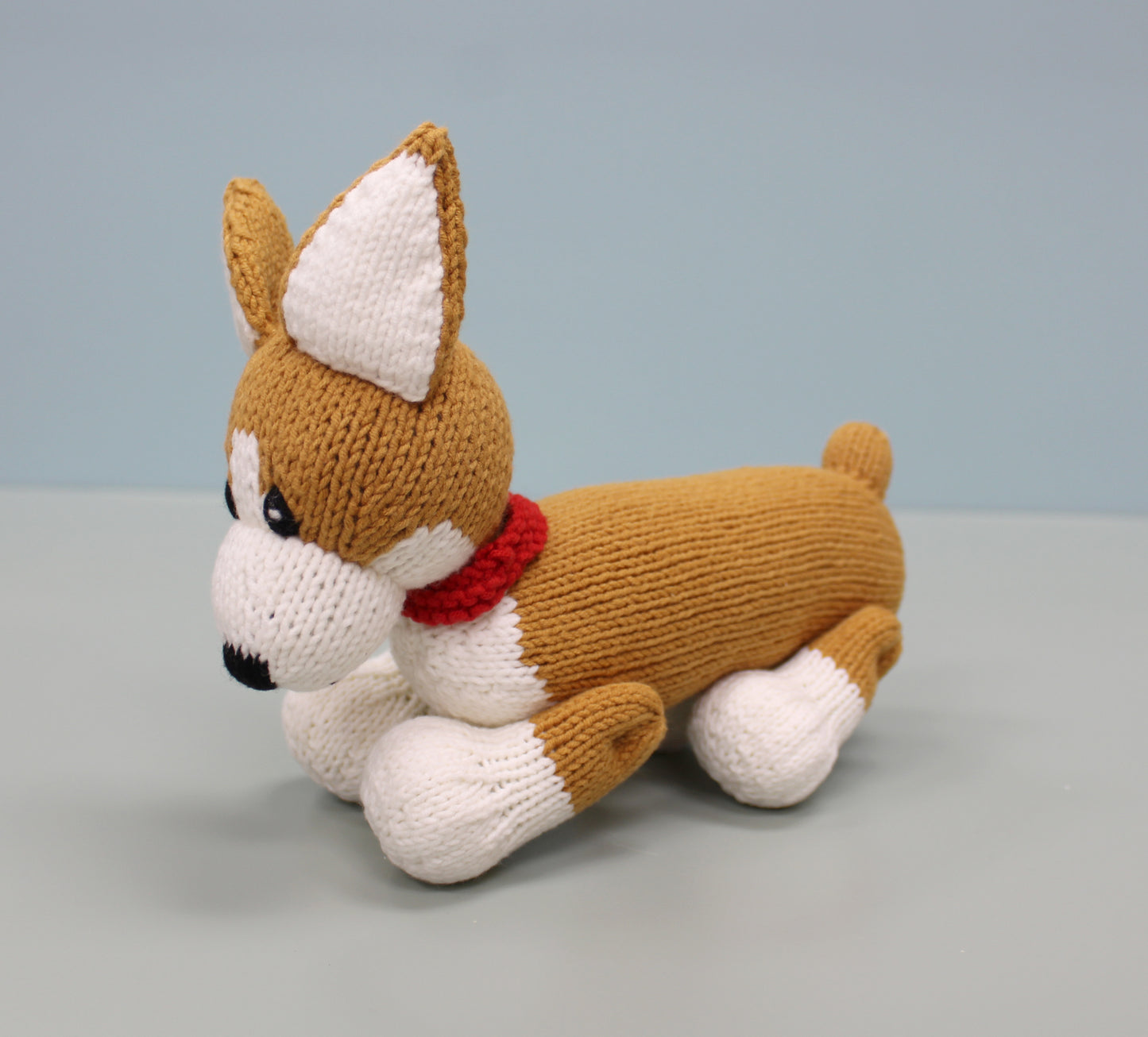 Handmade knitted corgi plush with brown and white fur, a red collar, and perky ears, lying down.