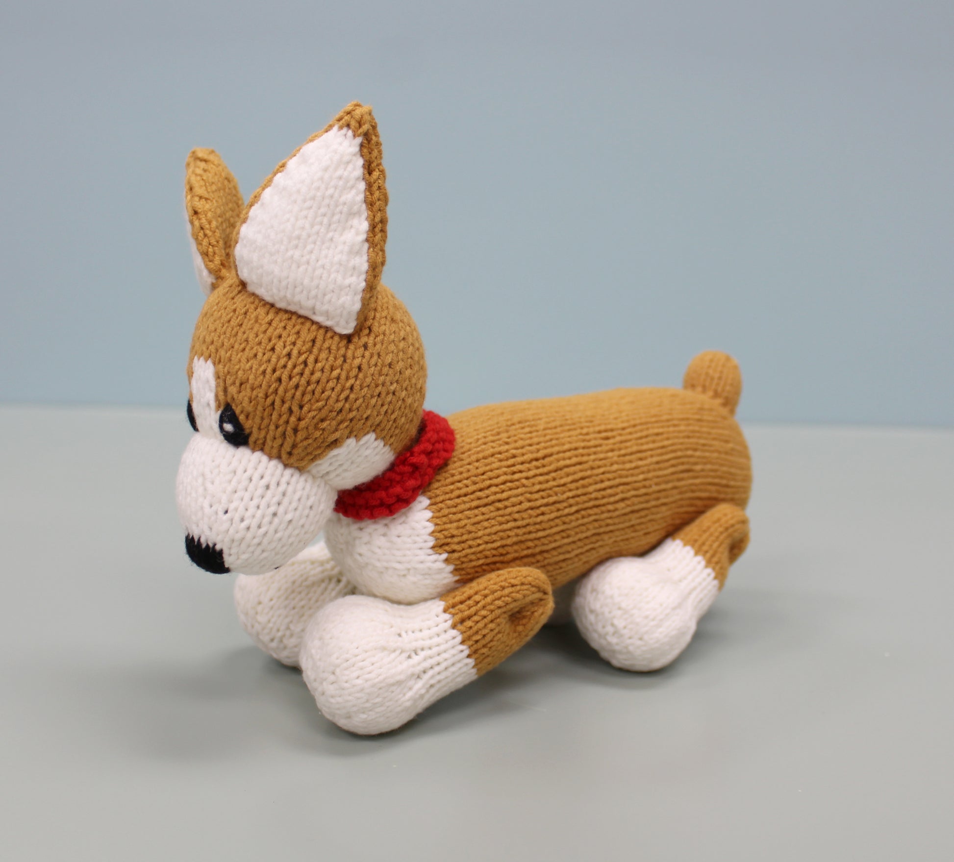 Handmade knitted corgi plush with brown and white fur, a red collar, and perky ears, lying down.