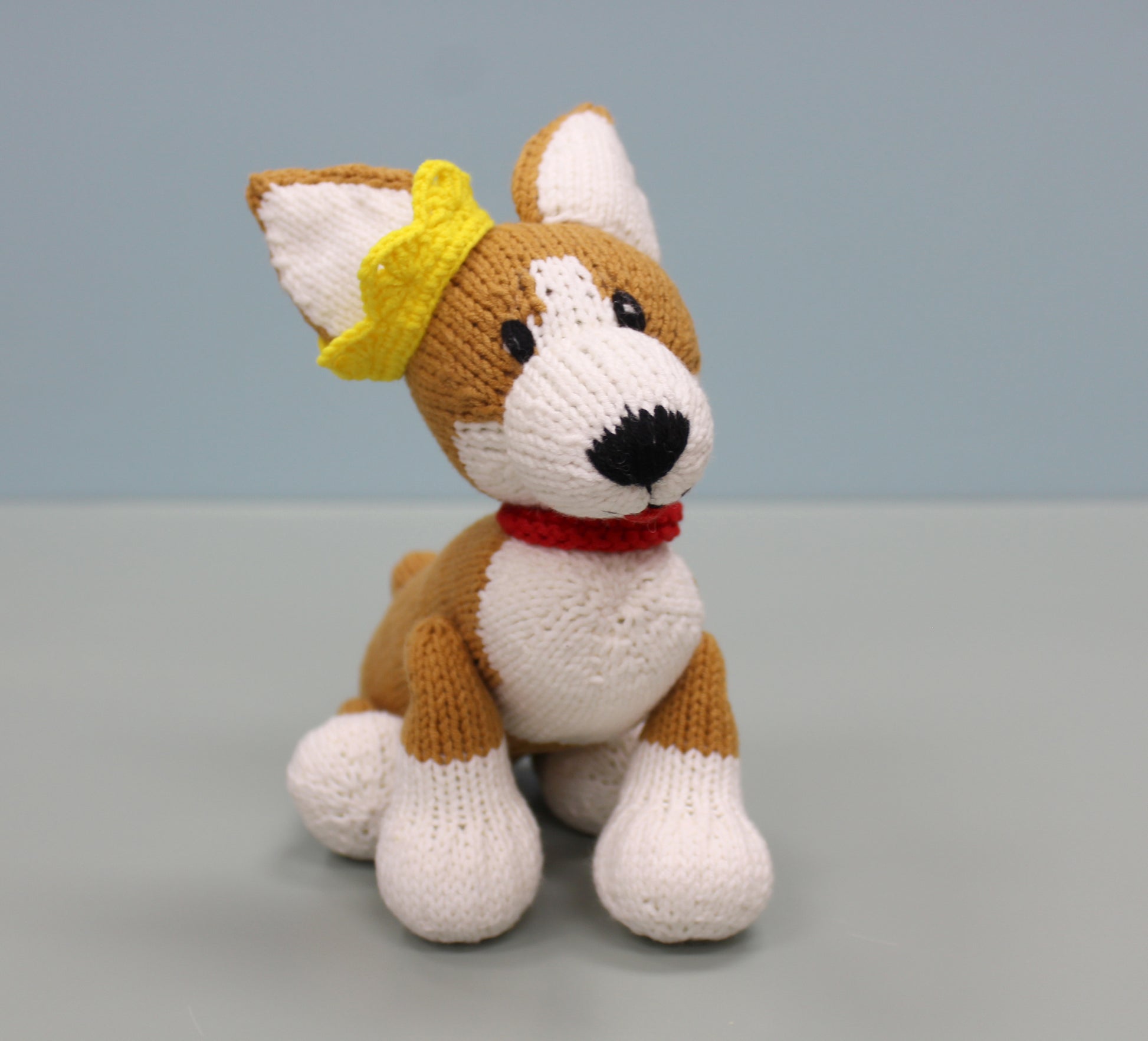 Brown and white handmade knitted corgi plush wearing a yellow bow and red collar.