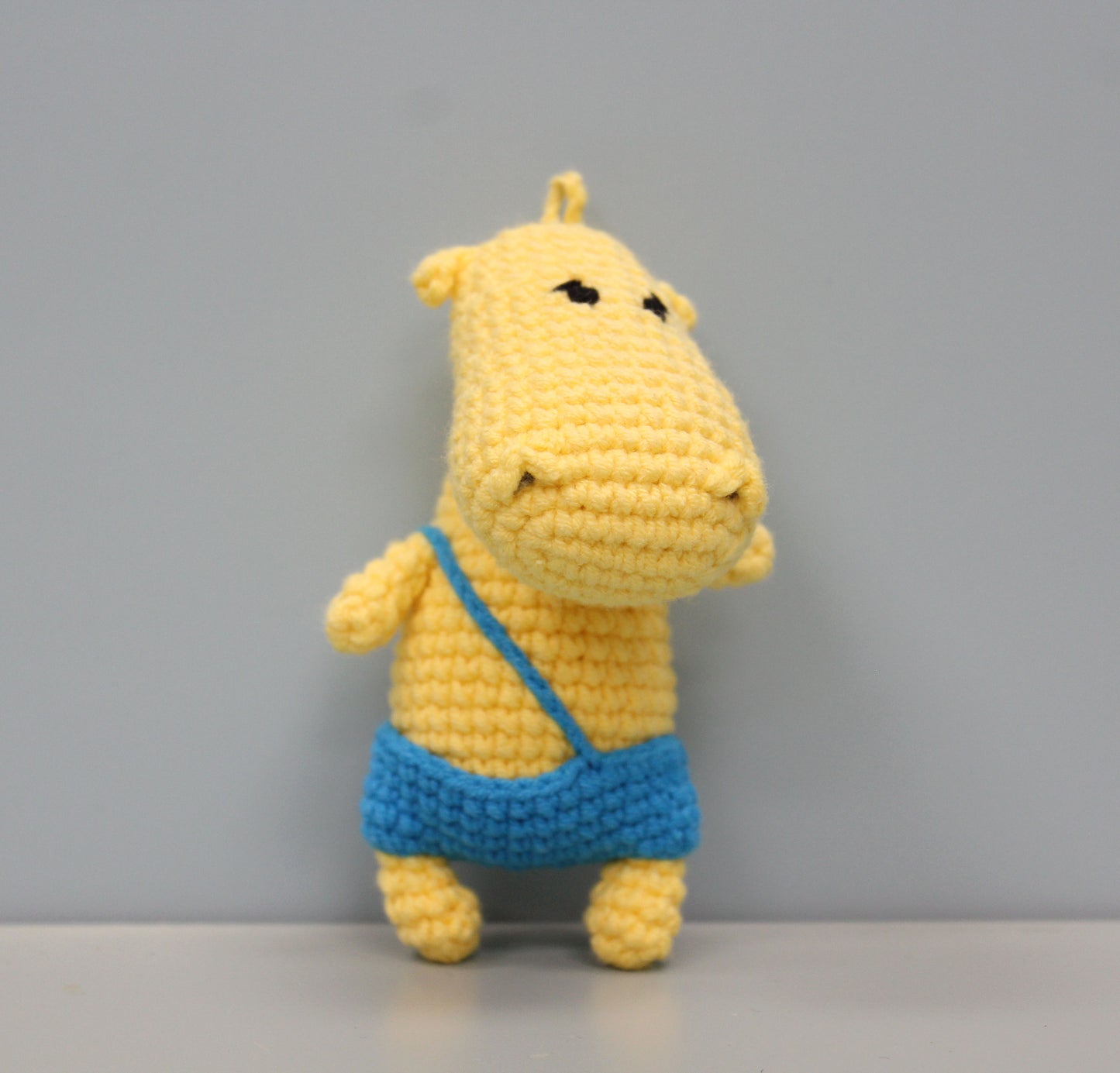 Handmade yellow crochet hippo keychain with blue shorts and a loop for hanging.