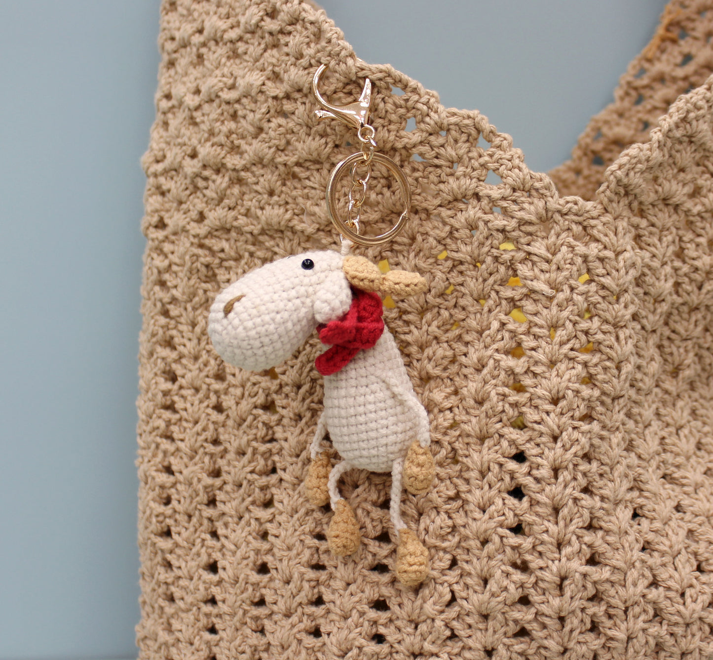 Handmade crochet moose keychain with a red scarf and gold clasp, displayed on a woven bag.