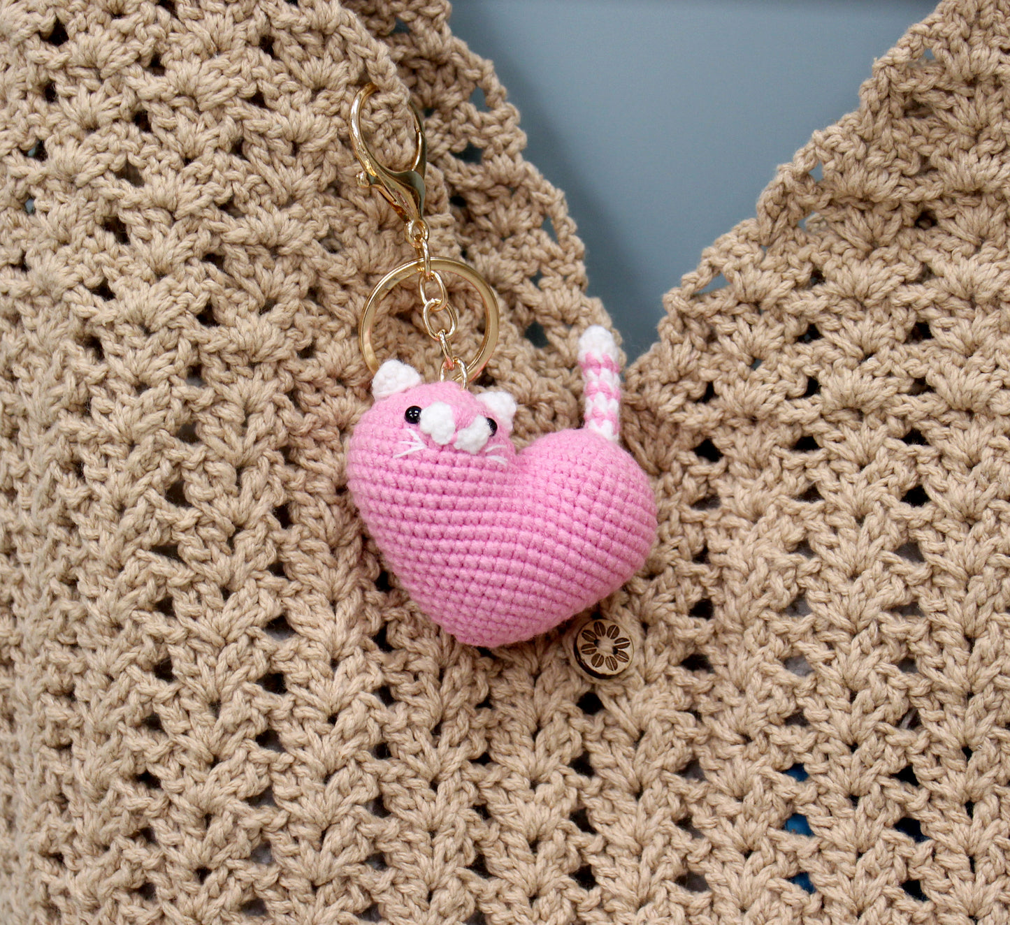Pink crocheted heart-shaped cat keychain with white accents and a gold ring, attached to a cozy knit sweater.